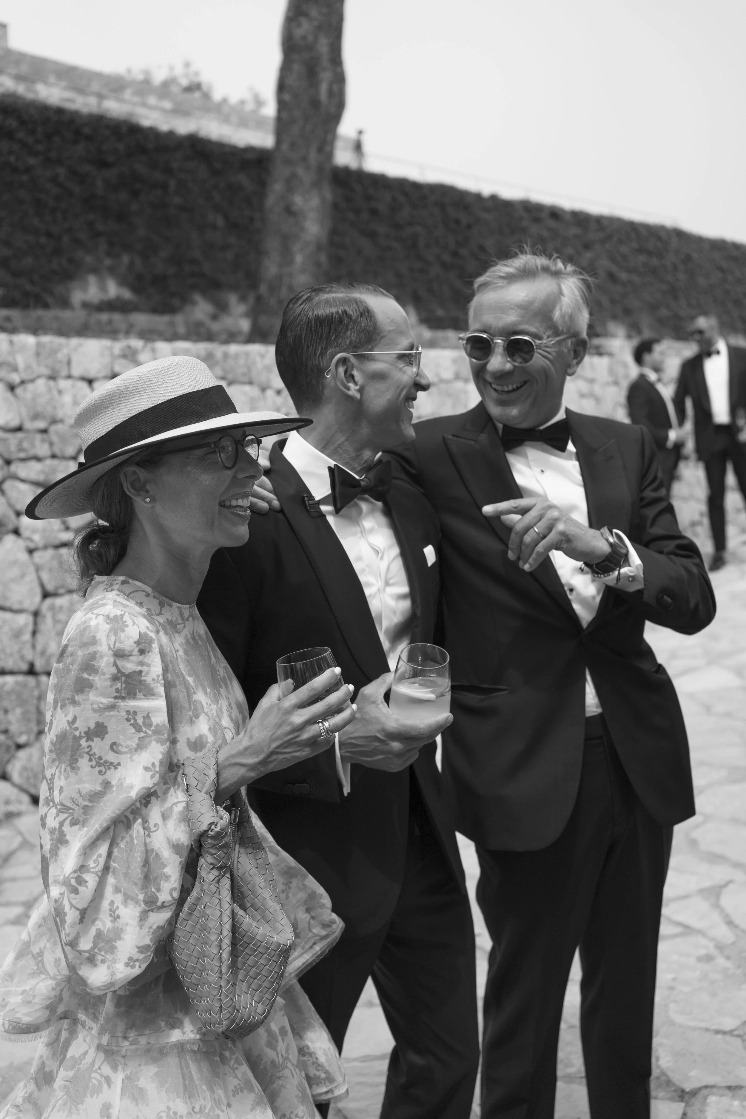 Three elegantly dressed people smiling and talking at an outdoor event, with two men in tuxedos and a woman wearing a floral dress and sunhat, holding drinks.