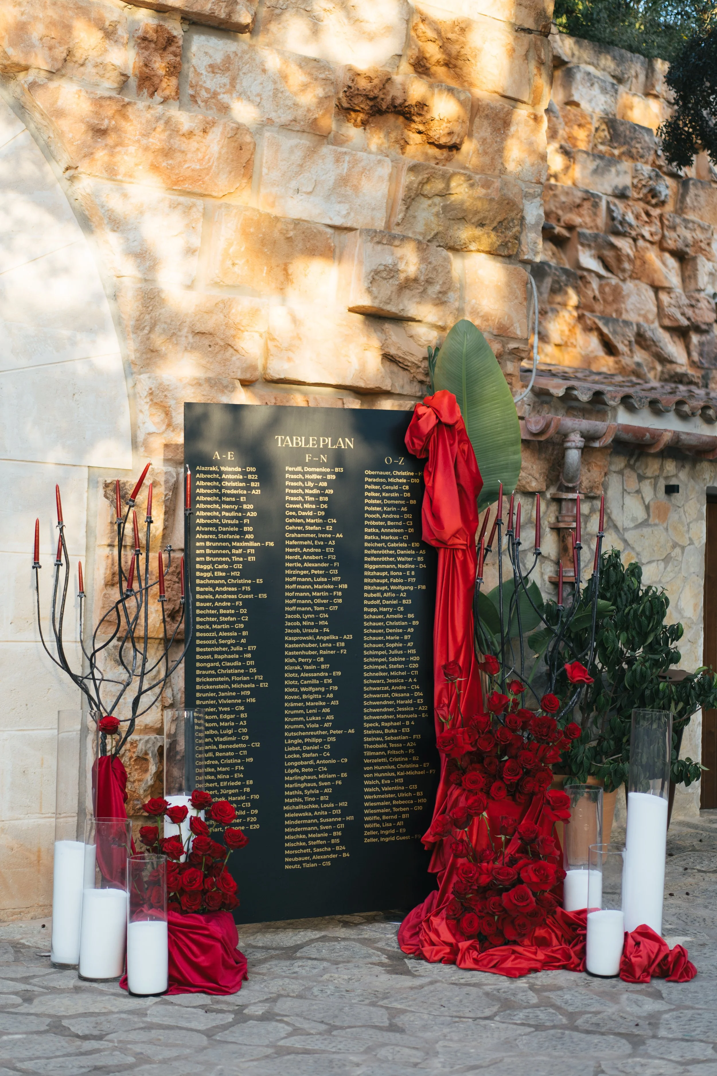A black wedding reception seating chart on a stand displayed outdoors, surrounded by red roses, candles, and floral arrangements, against a stone wall.