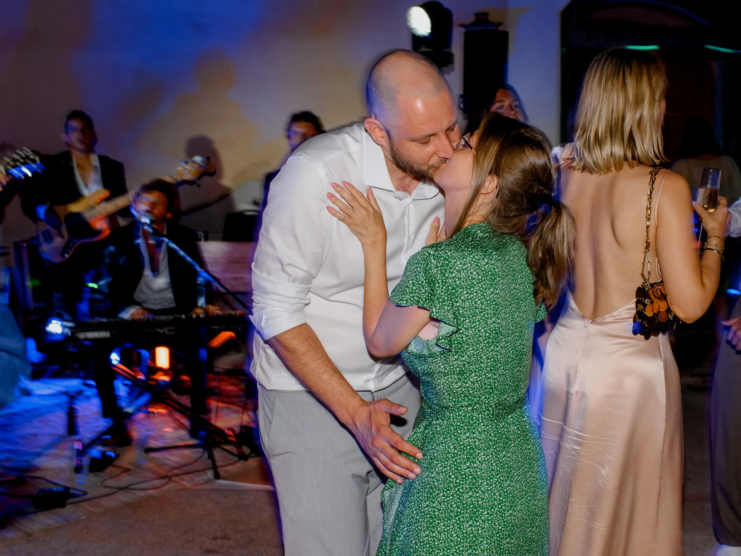A couple kissing at a party with a band playing in the background. The man is bald and wearing a white shirt, while the woman has long brown hair and glasses. A woman in a backless beige dress holding a drink is nearby.