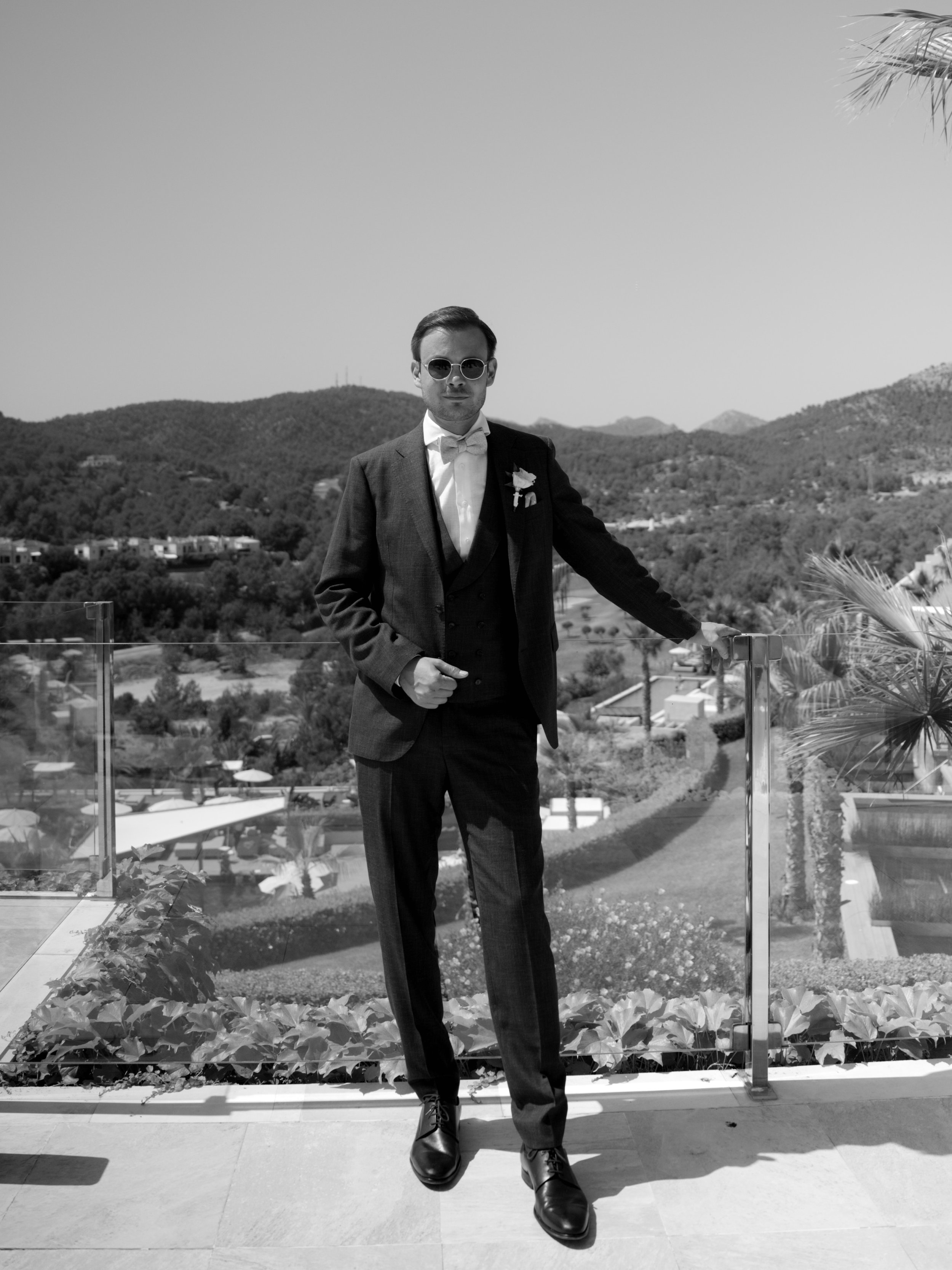 A man in a tuxedo with sunglasses standing outdoors on a balcony with mountains in the background.