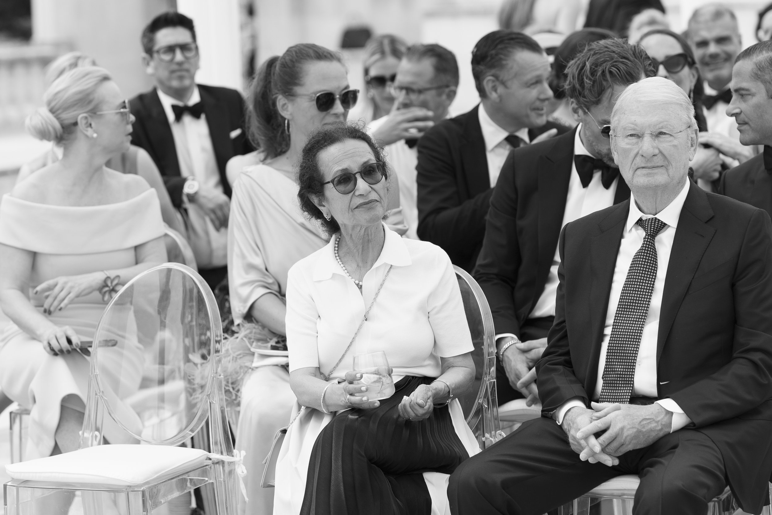 A black-and-white photo of a group of elegantly dressed people at an outdoor event, some wearing sunglasses, seated on clear chairs, with some standing and chatting in the background.