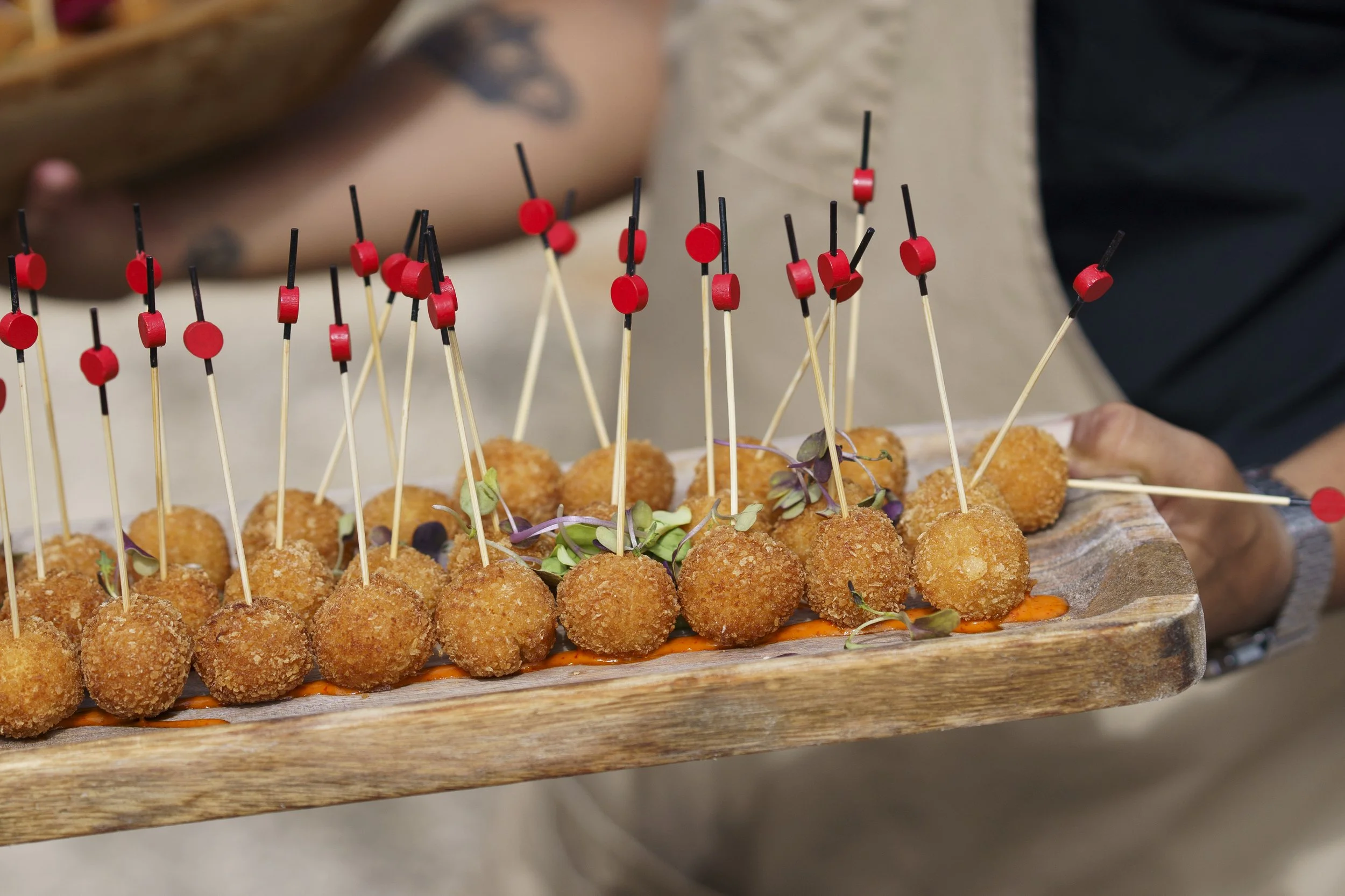 A wooden platter filled with fried appetizer balls on toothpicks, garnished with microgreens and sauce underneath.