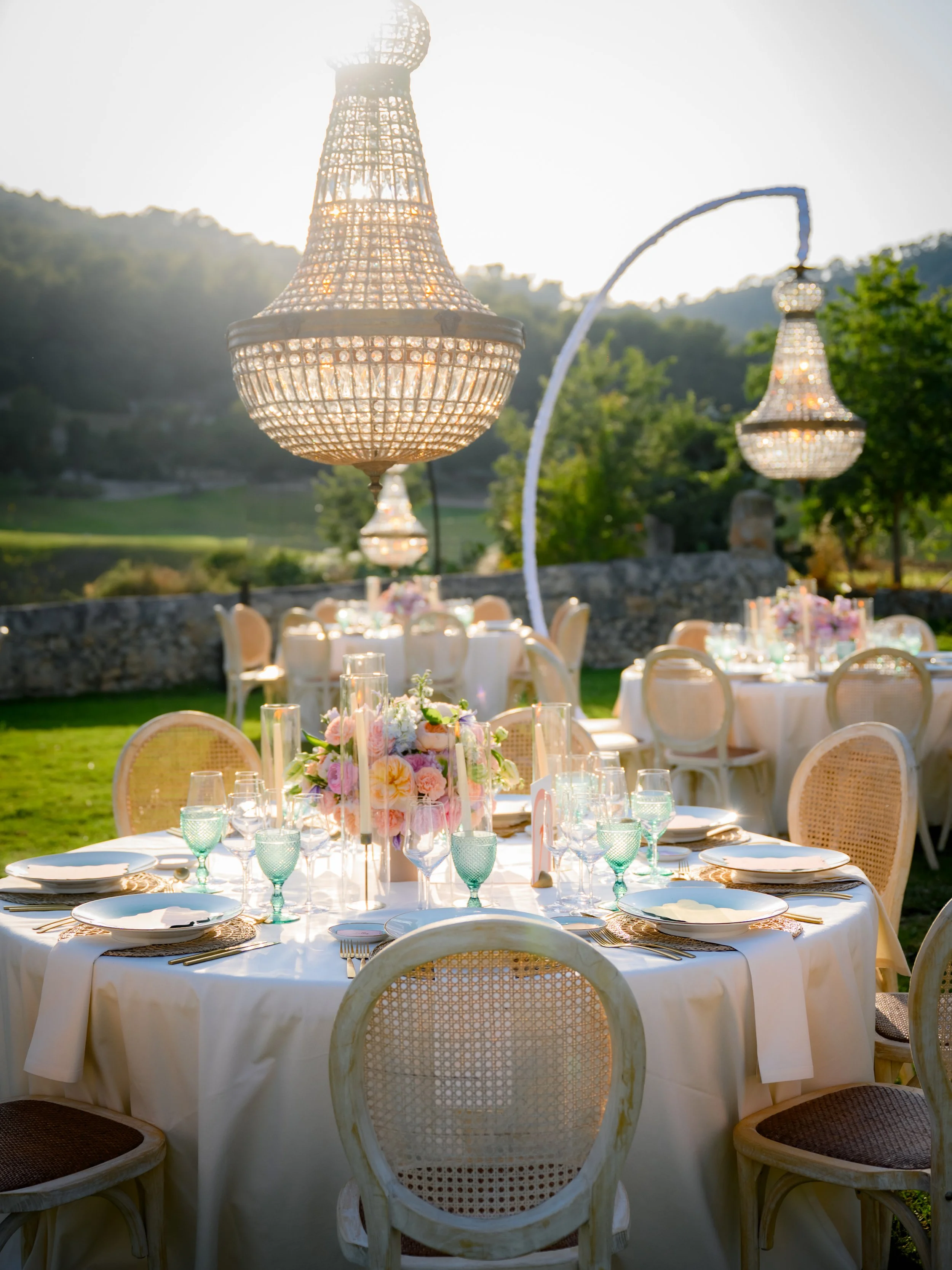 Outdoor elegantly decorated dining setup with round tables, pastel floral centerpieces, fine china, glassware, and chandeliers hanging overhead, set against a scenic landscape with greenery and hills.