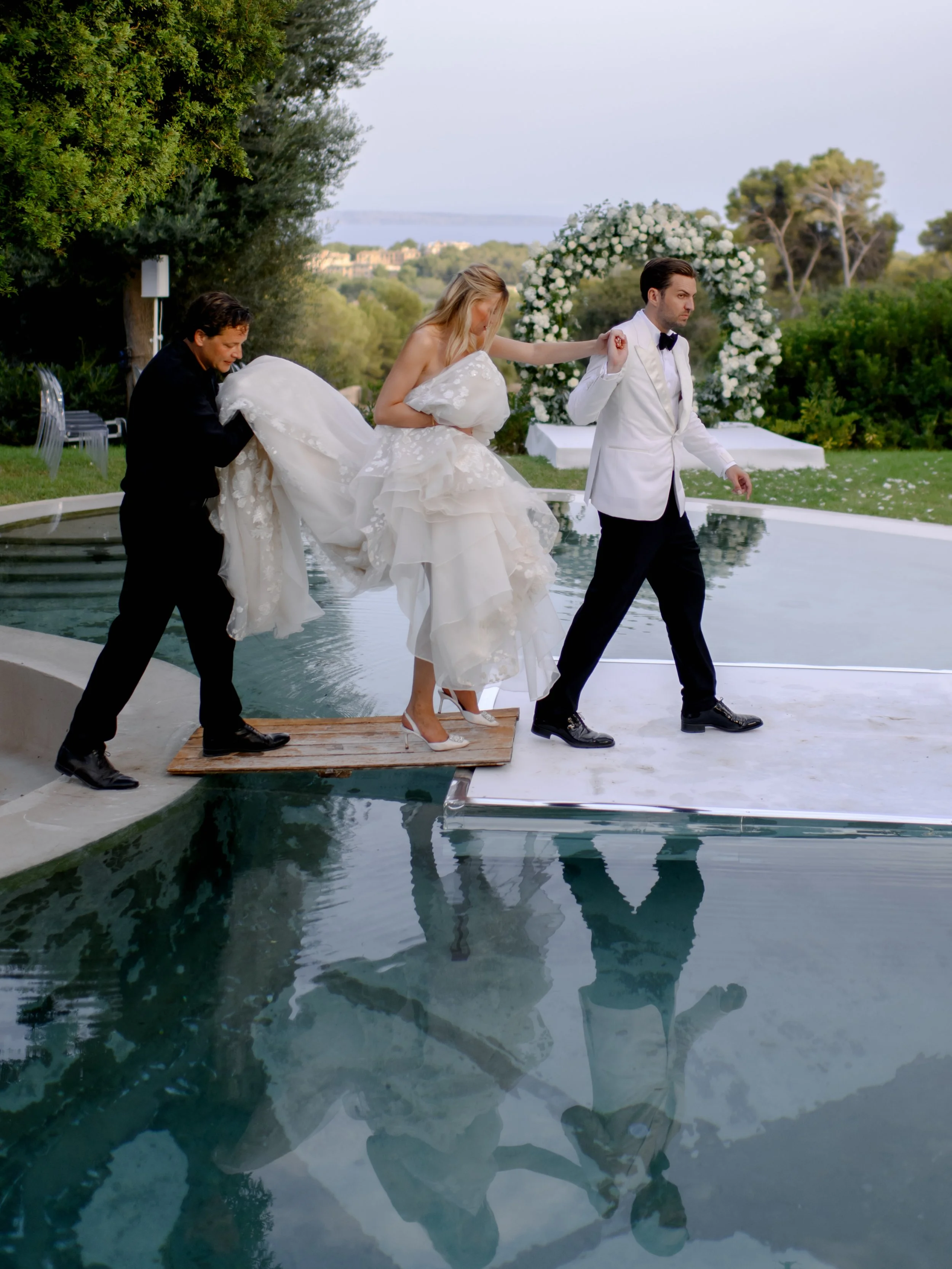 Bride being helped into a swimming pool by two men, one in a black suit and the other in a white tuxedo, at an outdoor wedding with greenery and a floral arch in the background.