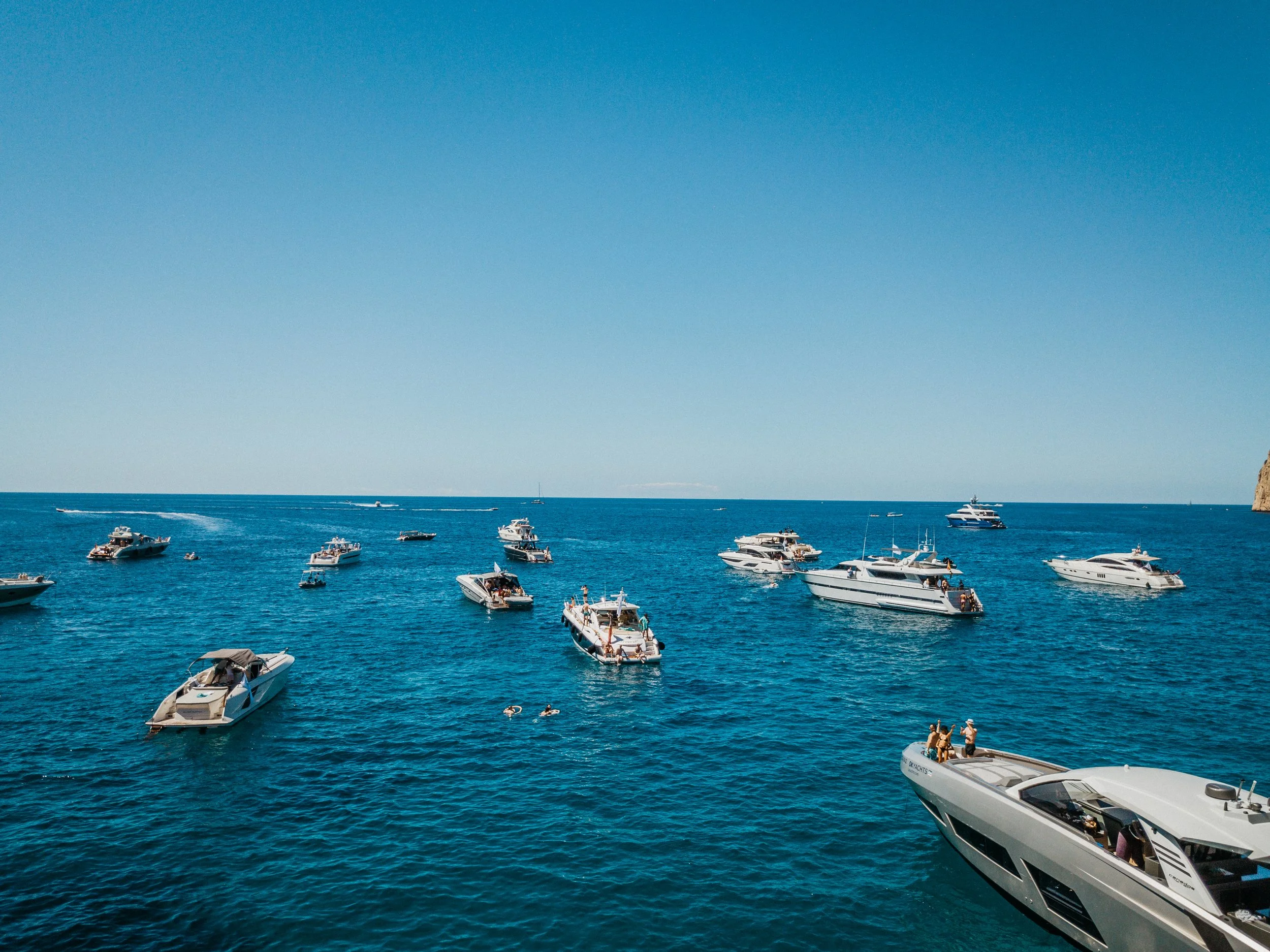 Multiple luxury yachts and boats floating on calm blue ocean waters under a clear blue sky.