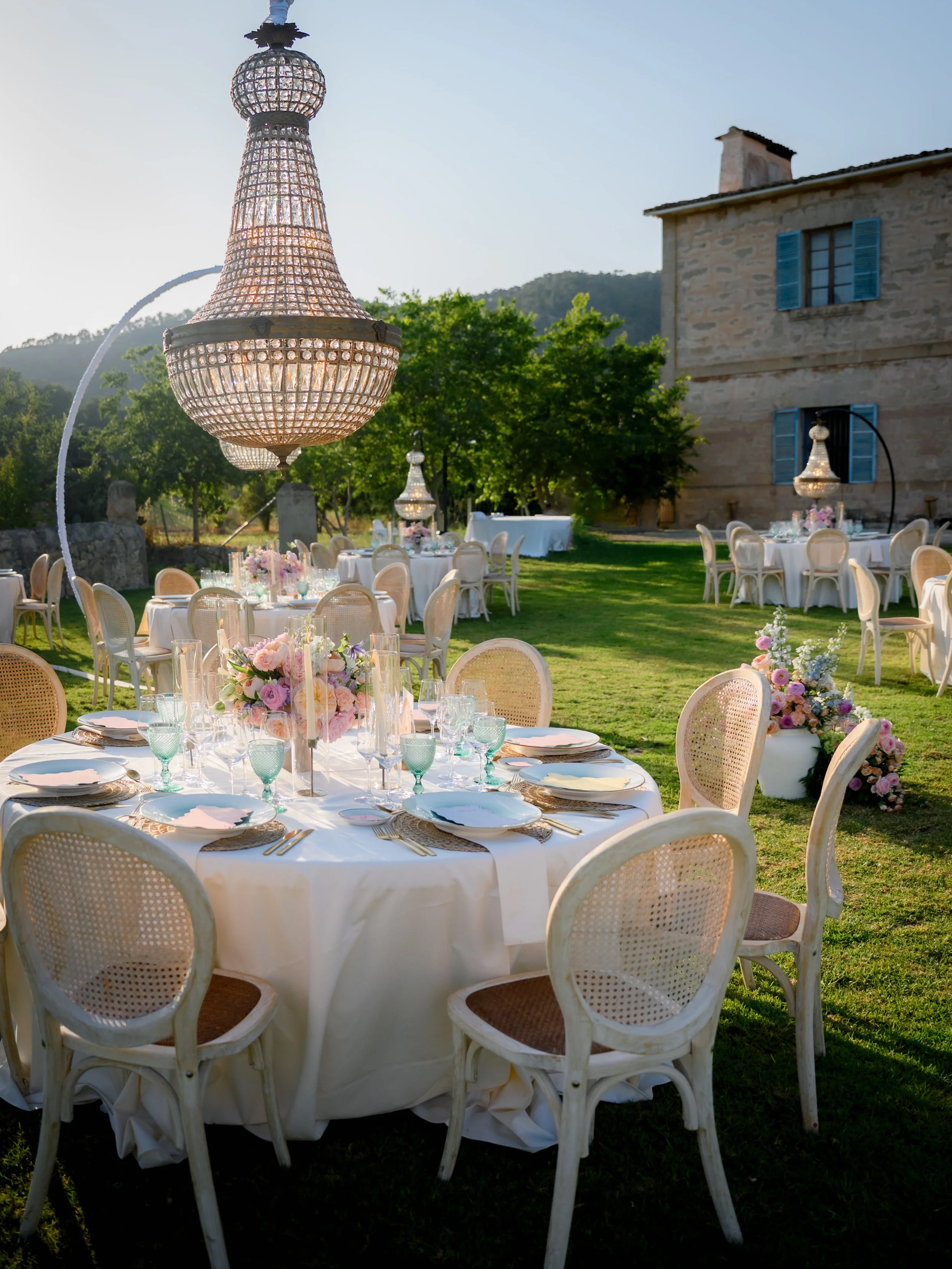 Outdoor wedding or event setup with round tables, floral centerpieces, chandeliers, and elegant chairs, in a grassy yard near a stone building with blue shutters.