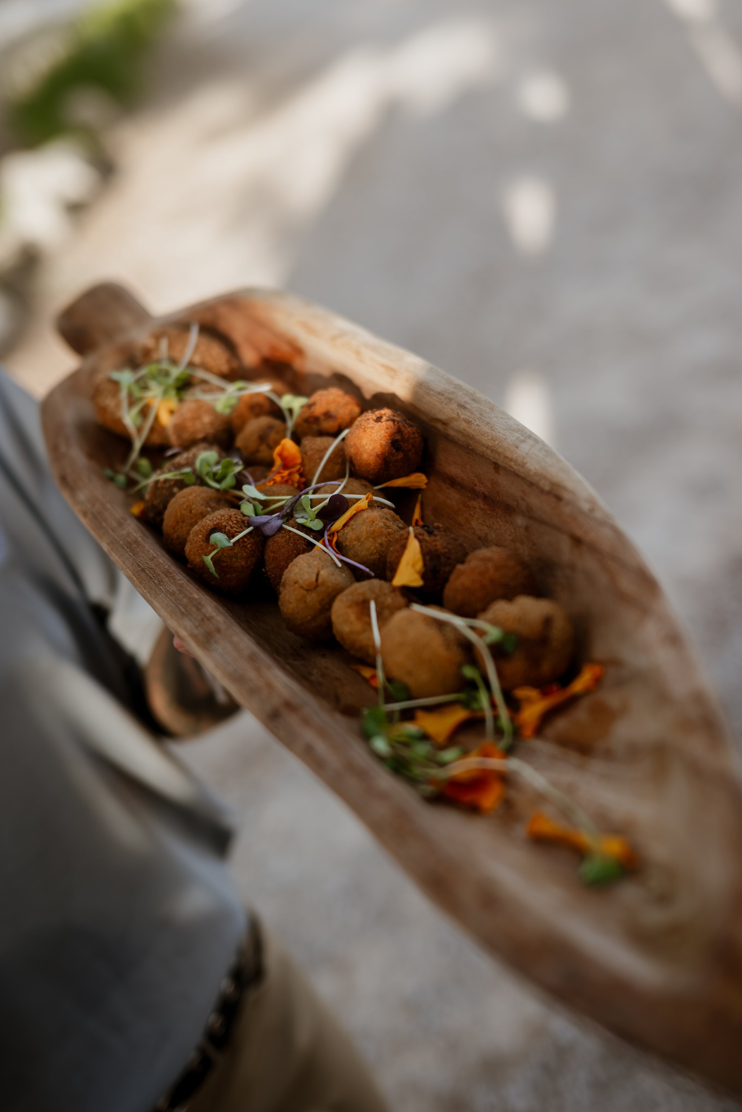 A wooden serving platter with fried falafel balls topped with edible flowers and microgreens.
