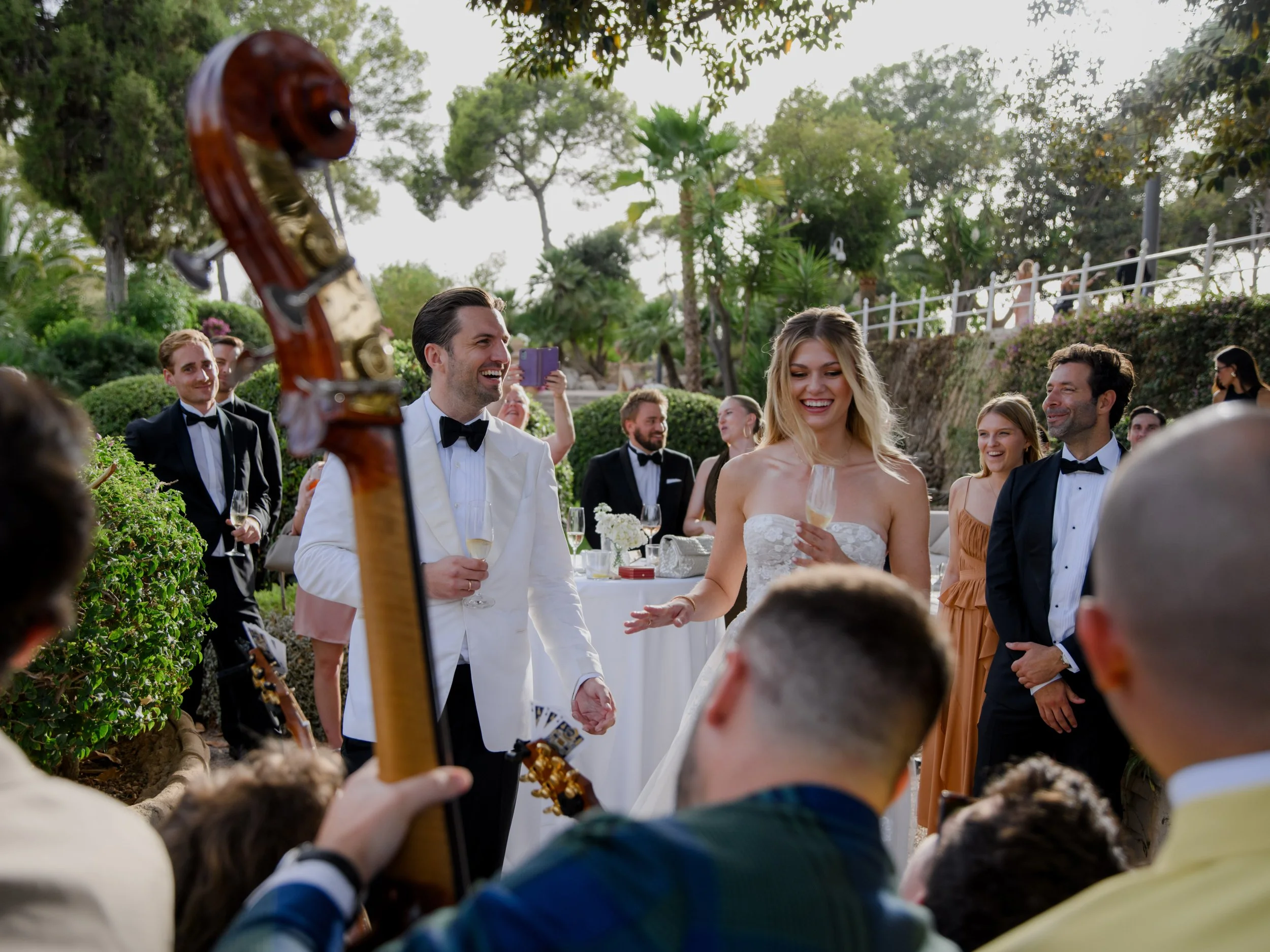 Wedding reception outdoors with guests dressed in tuxedos and dresses, bride in strapless white gown holding a champagne flute, groom in white tuxedo jacket laughing, a musician plays a double bass, guests joyful around the bride and groom.