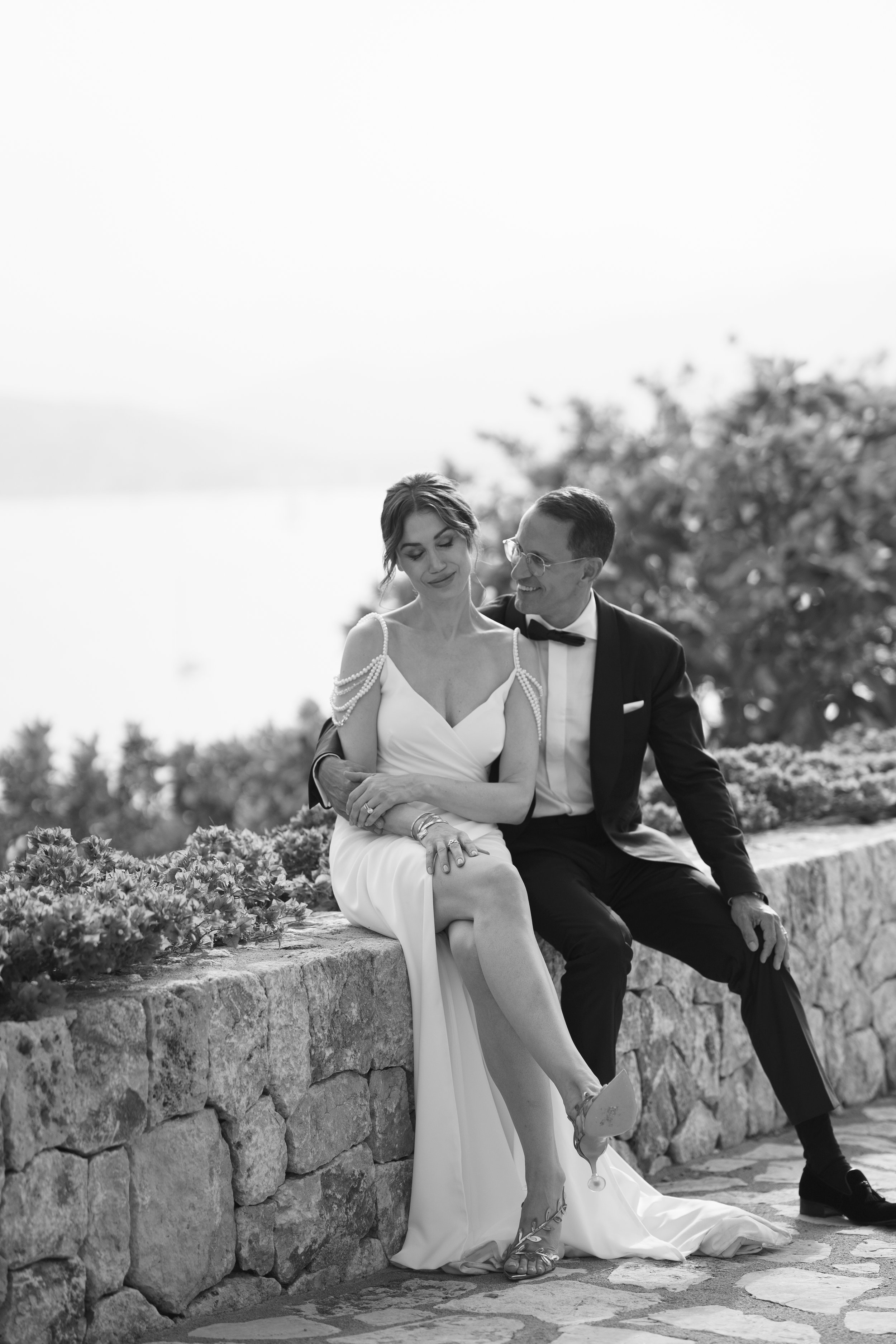 Black and white photograph of a bride and groom sitting on a stone wall outdoors, smiling and leaning close to each other.
