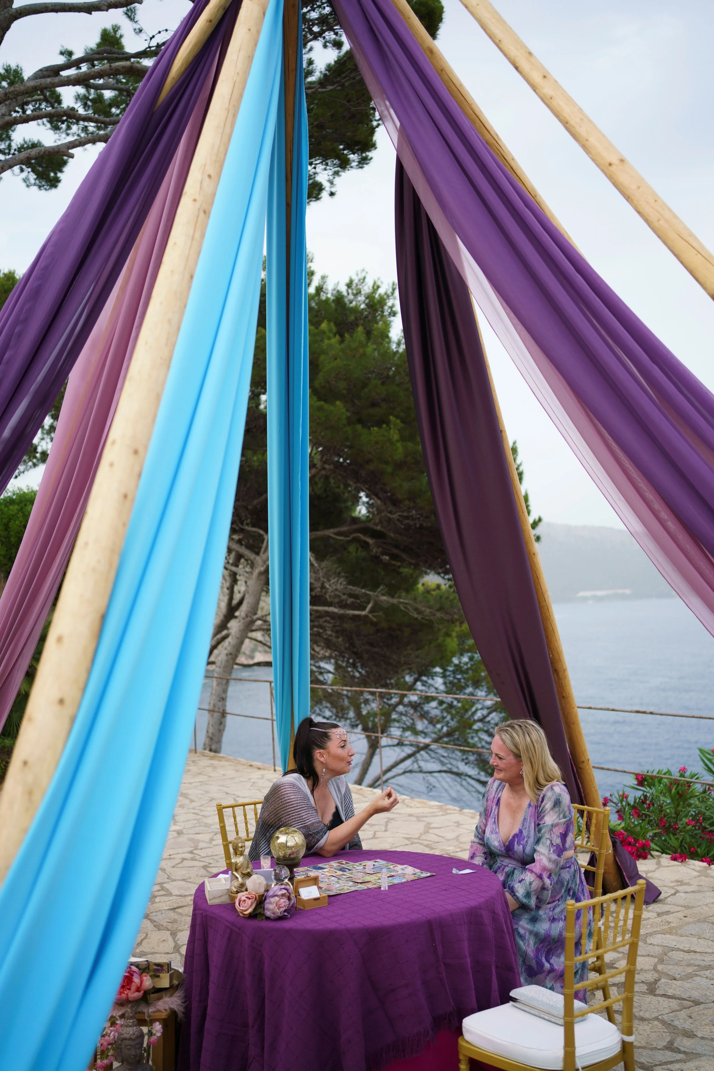 Two women sitting at a round table with a purple cloth, playing a game outdoors near the water, under colorful fabric drapes in purple, pink, light blue, and darker purple, with trees and water in the background.