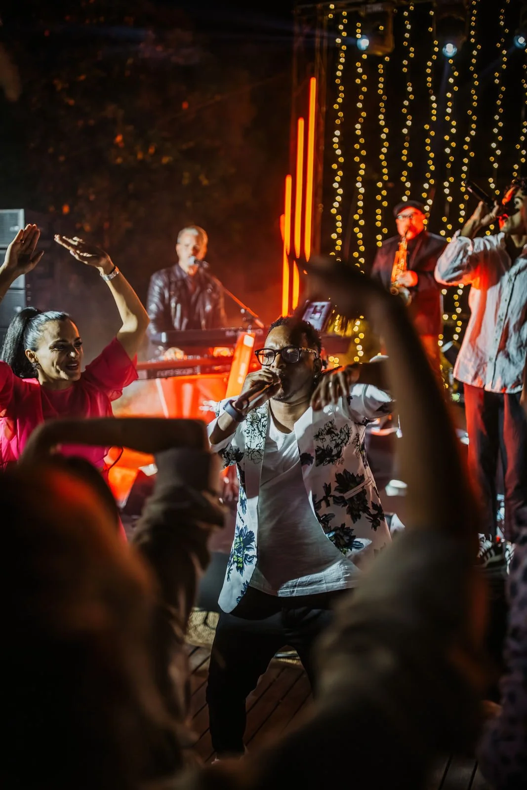People dancing and singing at a lively outdoor concert with a band performing on stage, lit by colorful stage lights and string lights in the background.