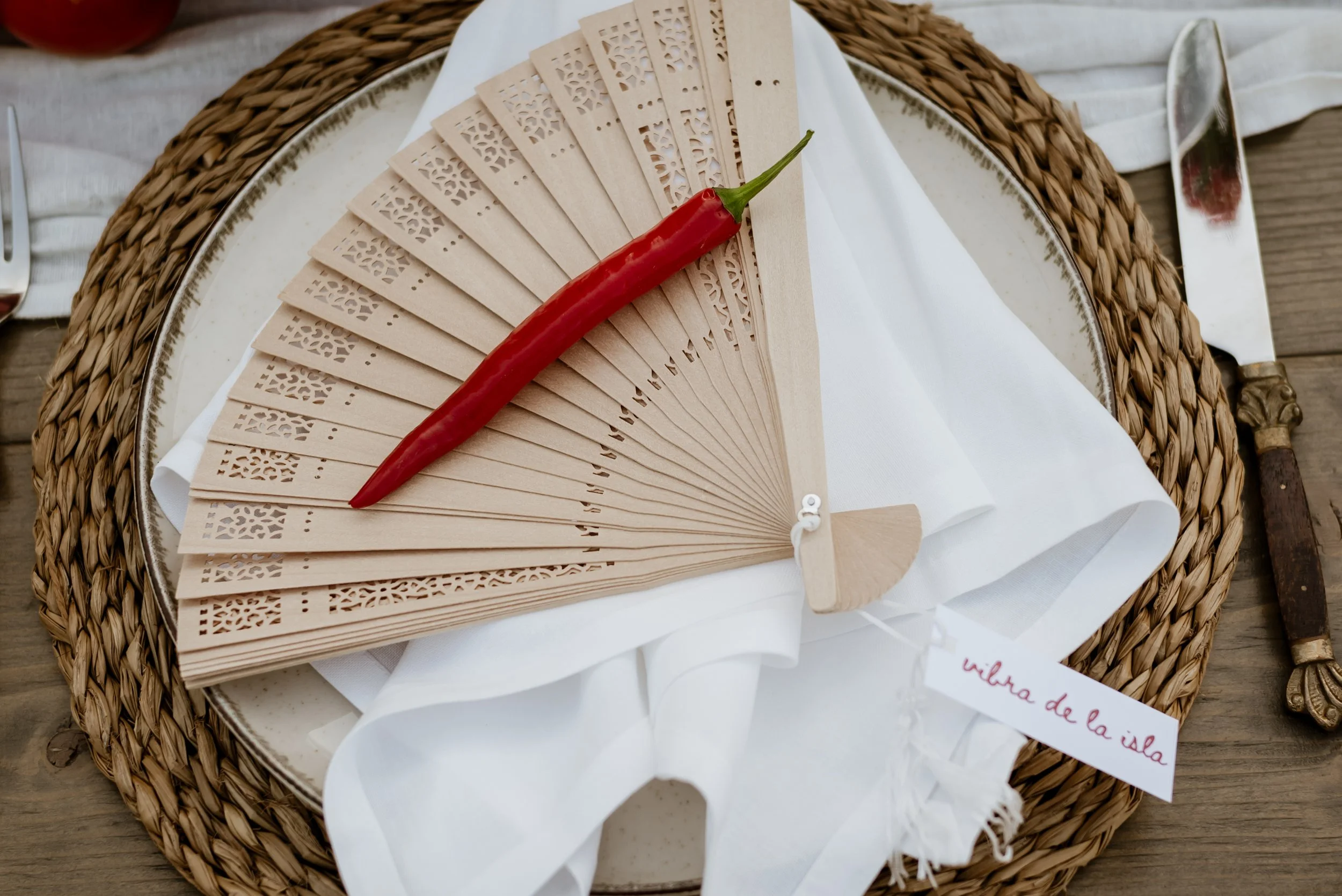 A decorative wooden hand fan with intricate cut-out patterns, a red chili pepper placed on top, on a white cloth and a woven placemat, with a small card reading 'villa de la isla'. A knife with an ornate handle and a fork are partially visible.