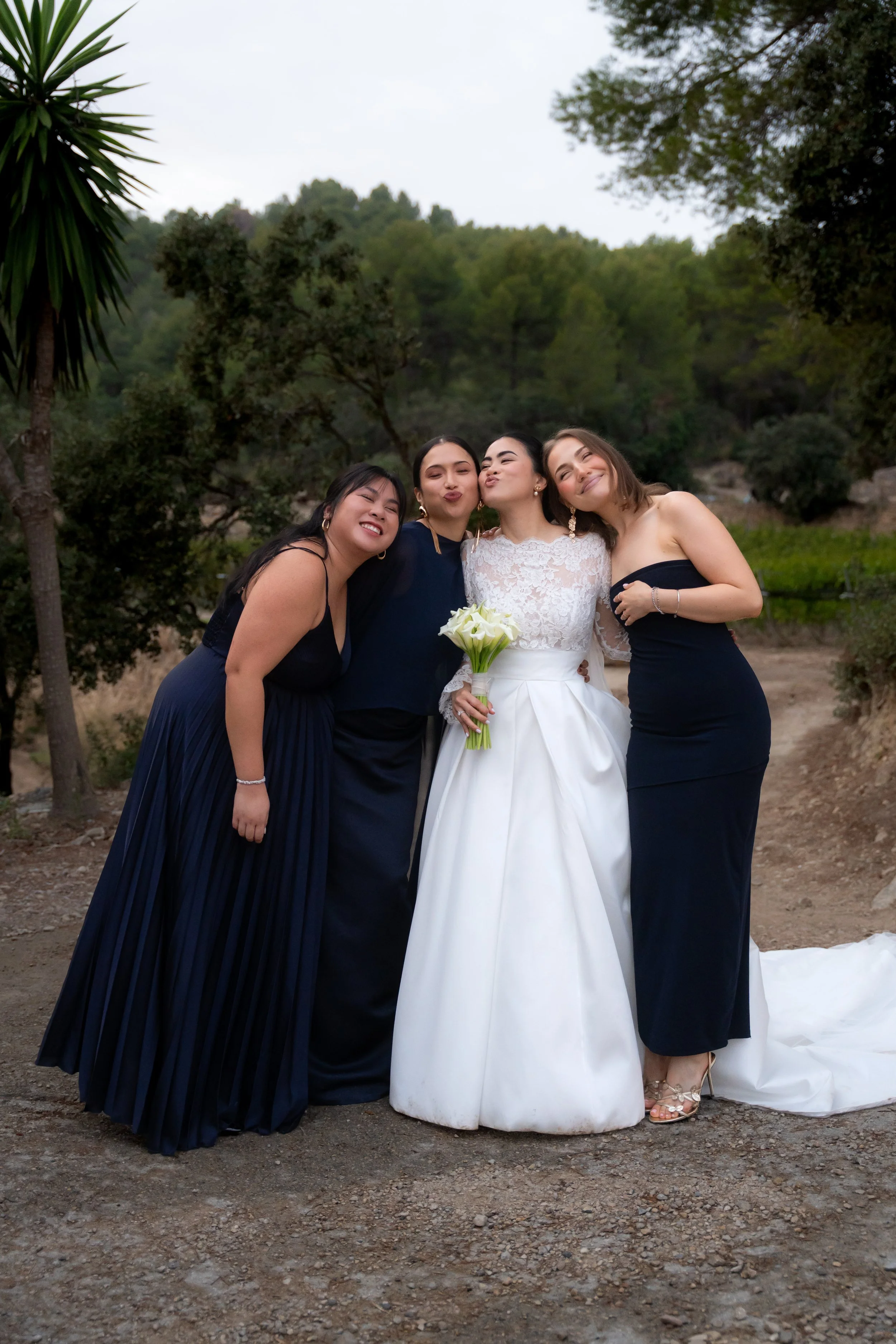 A bride and three women, possibly bridesmaids, standing outdoors on a dirt path with trees in the background. The bride is wearing a white wedding dress and holding a bouquet of white calla lilies. The women are dressed in dark dresses and are close 