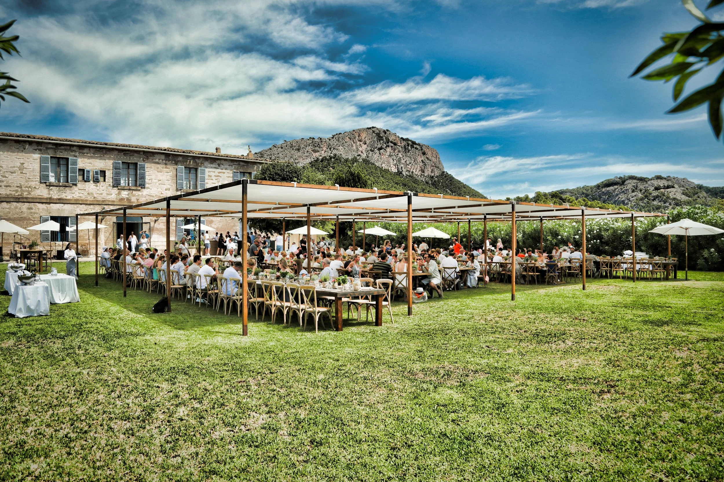 Outdoor wedding reception with numerous guests seated at tables under a canopy on a grassy lawn, with a historic stone building, trees, and mountains in the background on a sunny day.