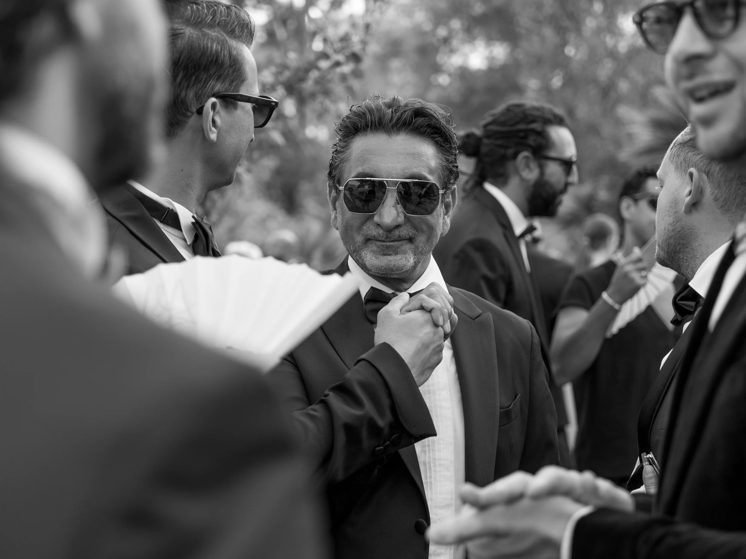 Black and white photograph of a group of men in formal tuxedos at an outdoor event, socializing and smiling, with one man wearing sunglasses in the center.