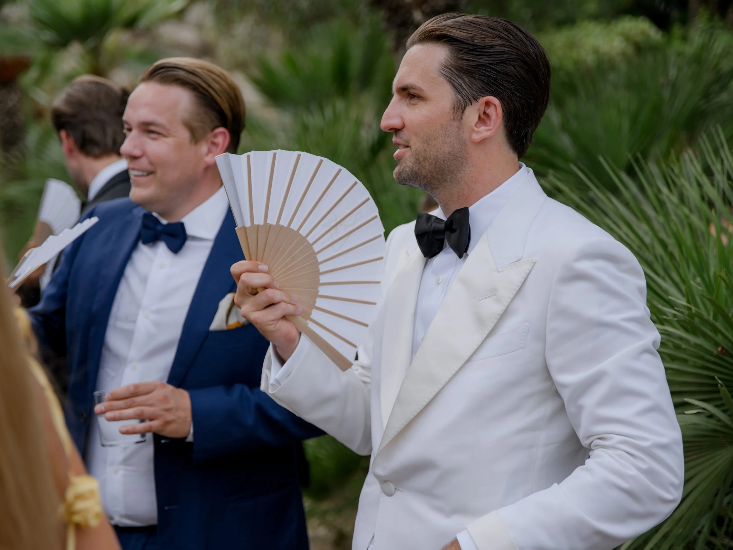 Several men in formal suits and tuxedos at an outdoor event with greenery, holding folding fans.