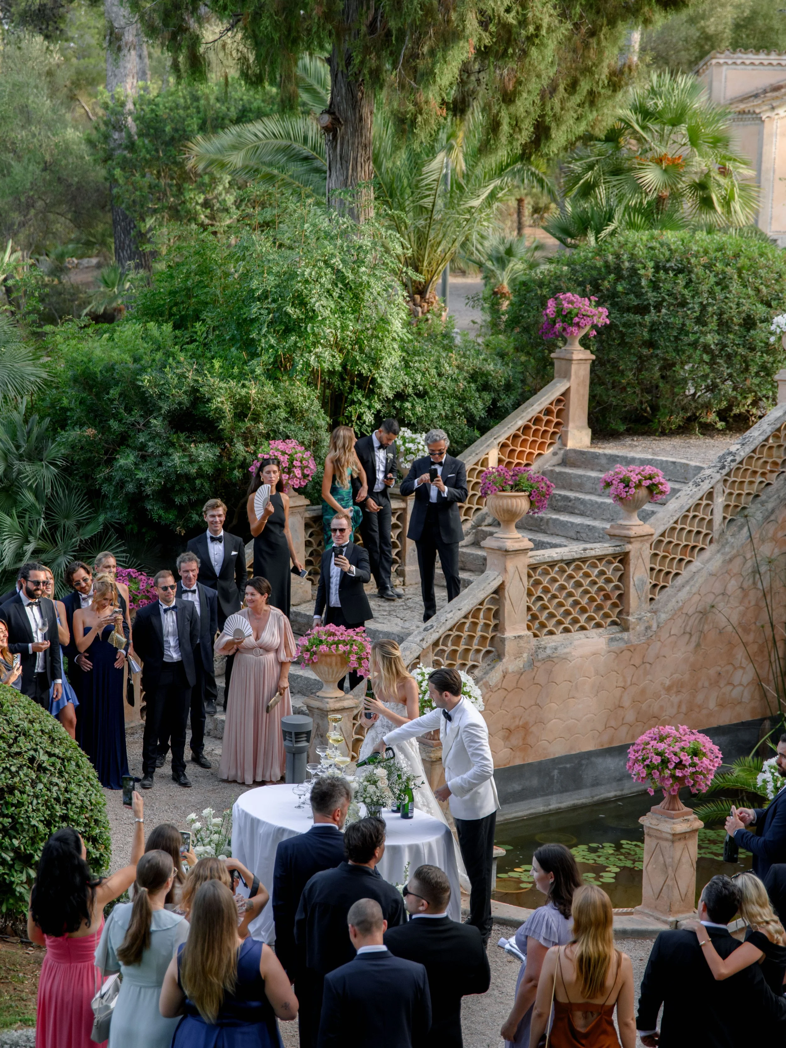 A wedding ceremony taking place outdoors on a tiered stone staircase, surrounded by lush green plants and pink flowers, with a bride and groom at a small round table, and guests in formal attire watching.