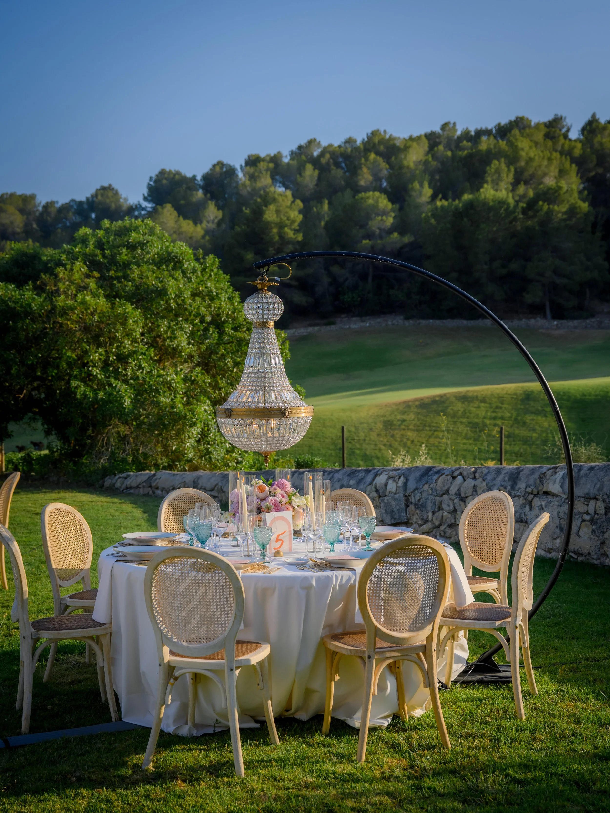 Outdoor dining table set with elegant glassware and floral centerpiece, under a large chandelier, in a lush green garden with a stone wall and rolling hills in the background.
