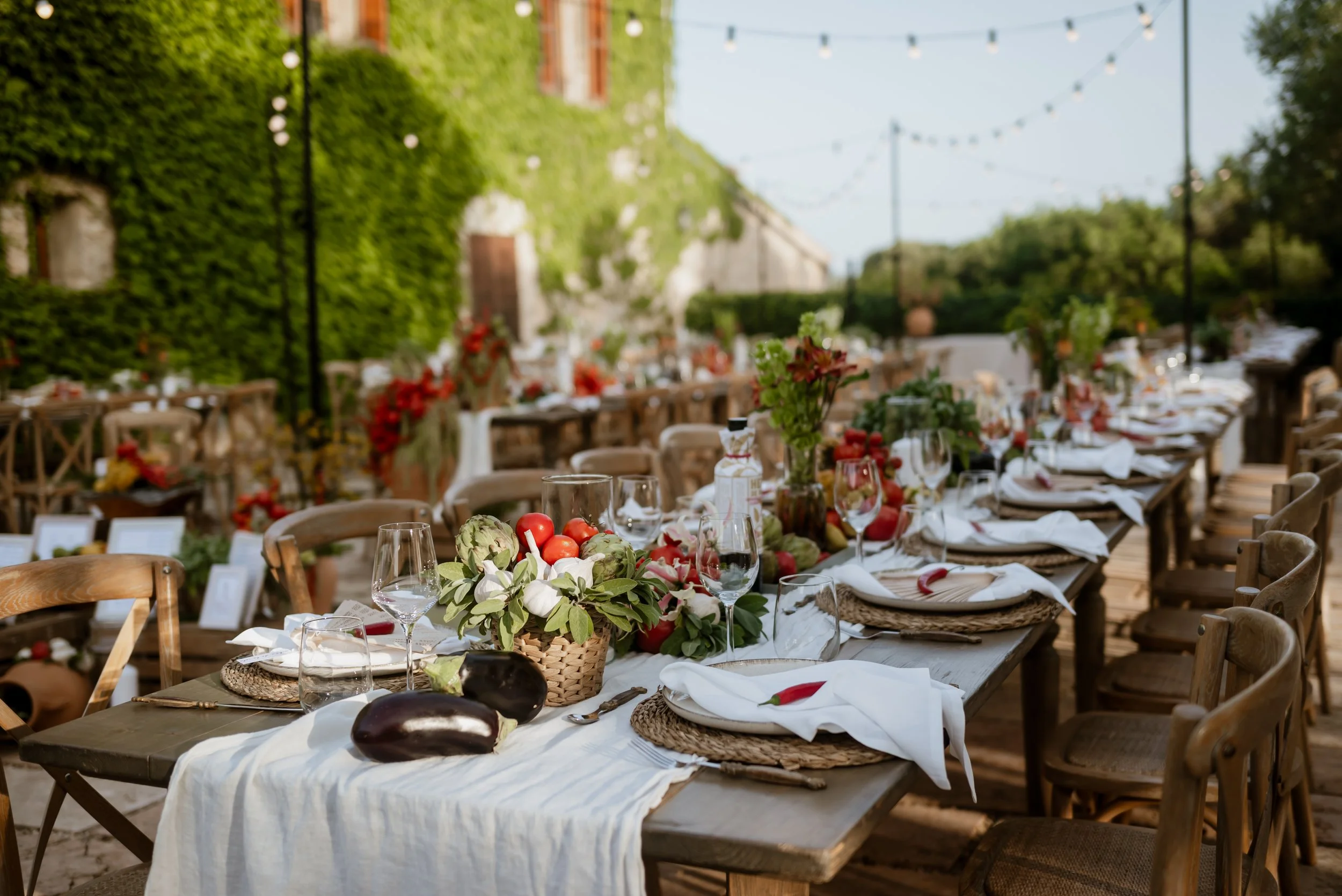 Outdoor dining table set for a celebration with floral centerpieces, plates, glasses, and utensils, decorated with vegetables and surrounded by wooden chairs, with string lights overhead and greenery in the background.