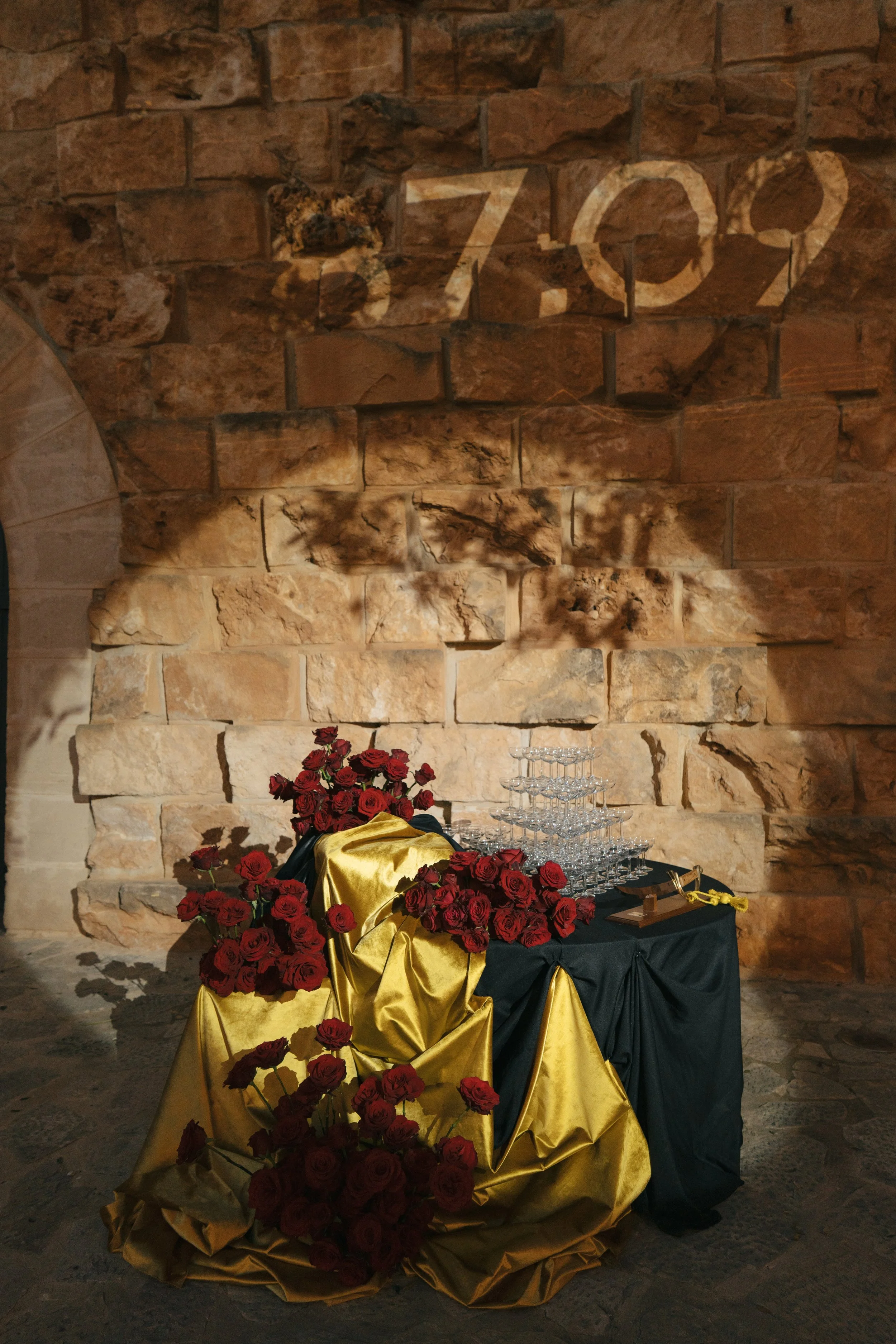 A table decorated with red roses, gold and black fabric, and stacked glasses against a stone wall with the word 'Zoz' spelled out on it.