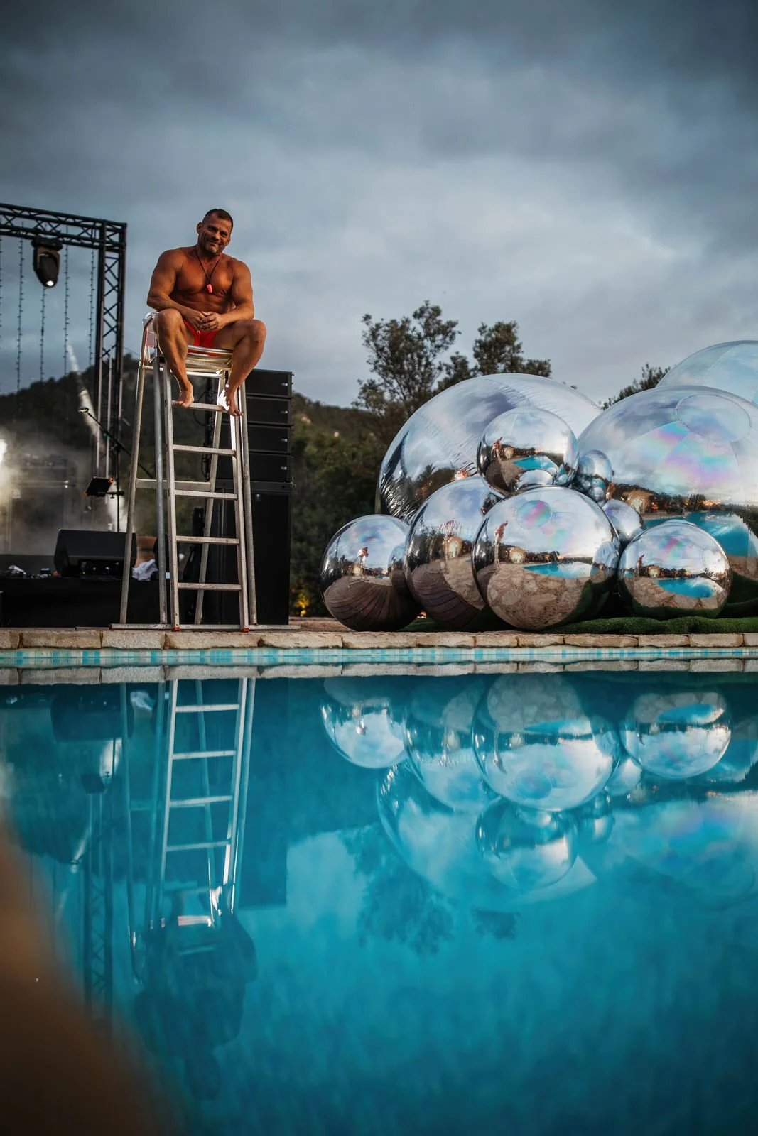 A man in red shorts sitting on a ladder at a poolside during twilight, with large reflective metallic spheres beside him and a stage with lighting equipment in the background.