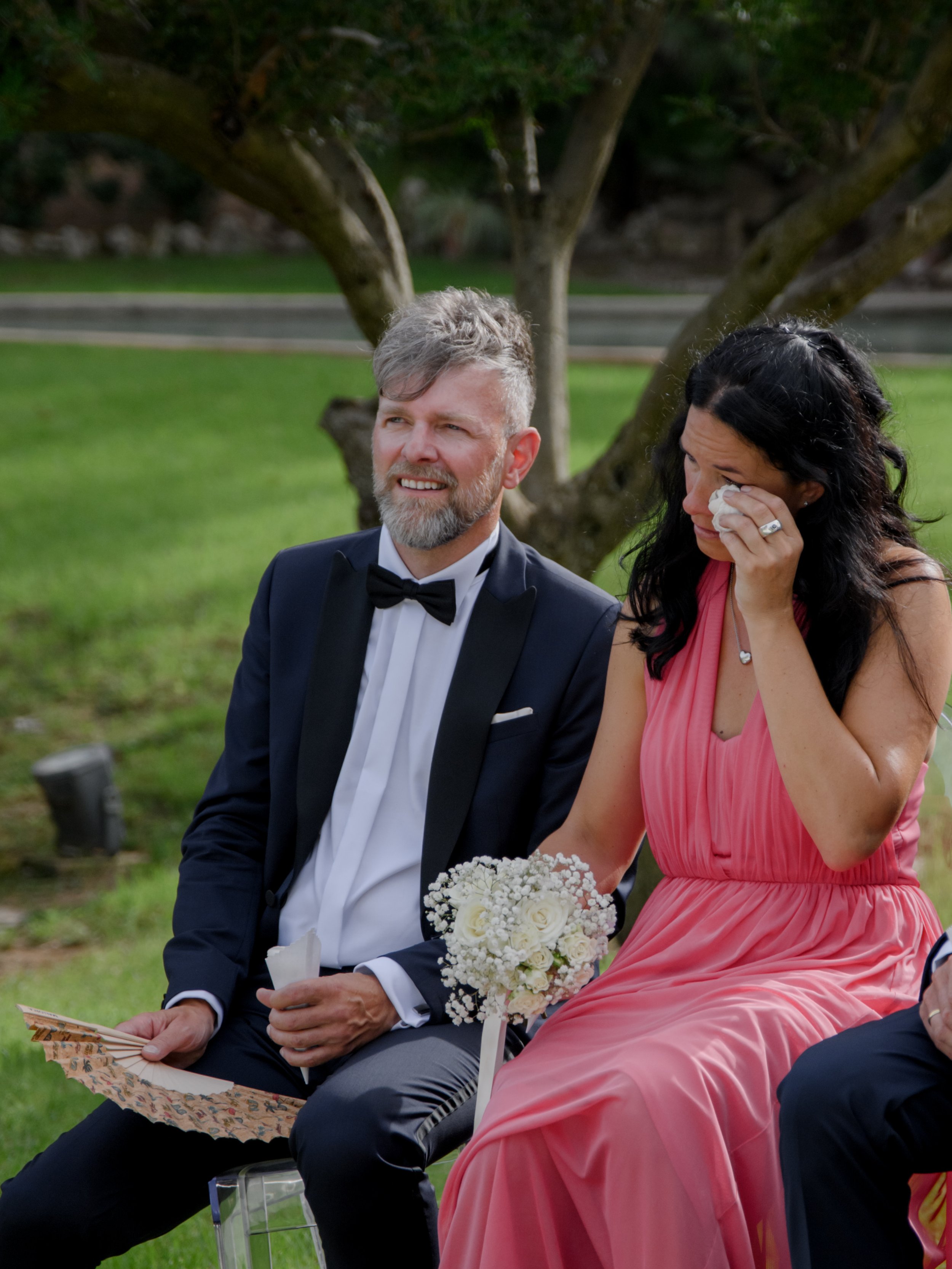 A man in a tuxedo and a woman in a pink dress sitting outdoors under a tree, with the woman wiping away tears and holding a bouquet of white flowers.