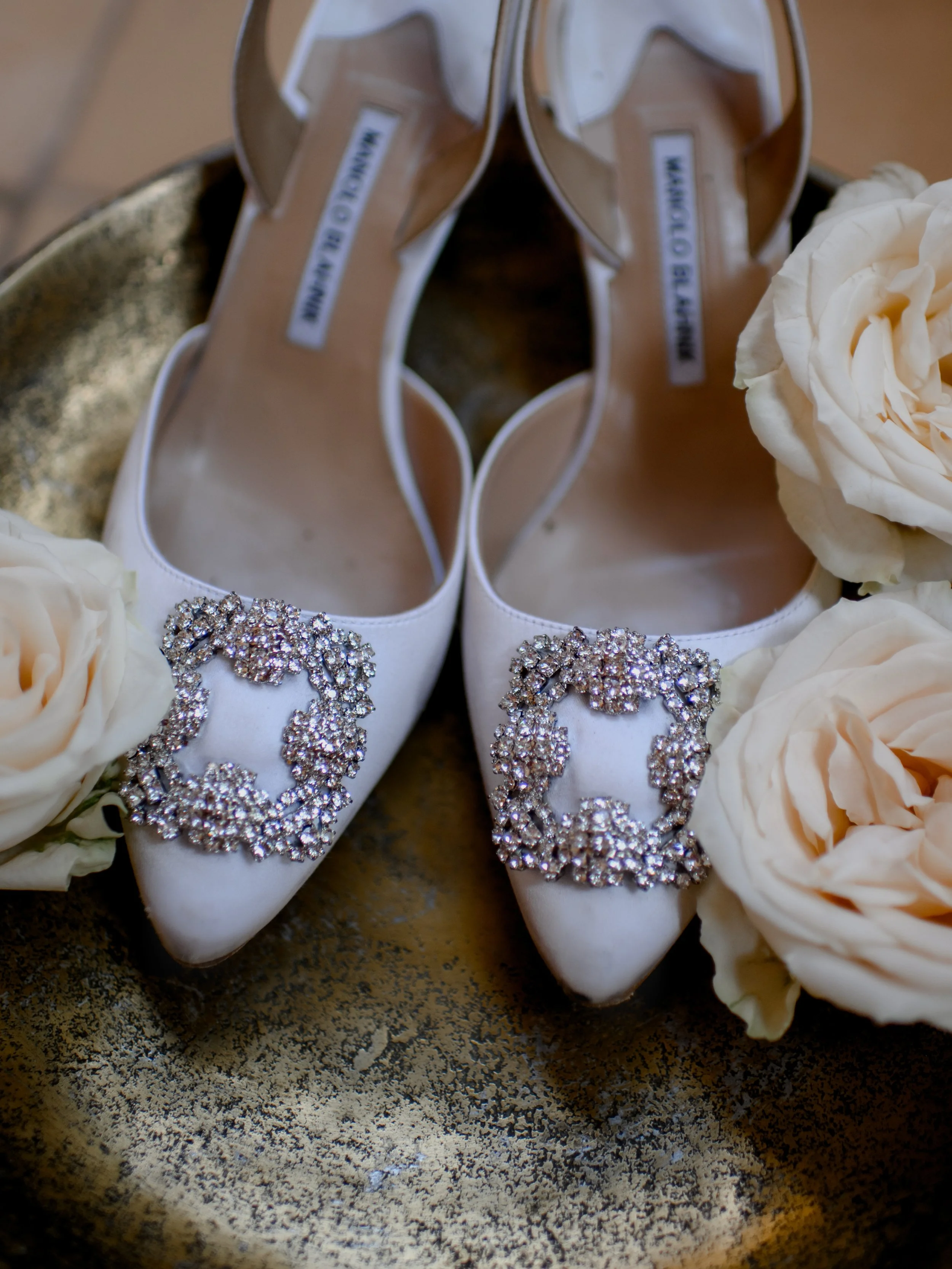 White high-heeled shoes with rhinestone embellishments on the toes, surrounded by white roses, placed on a gold-colored tray.