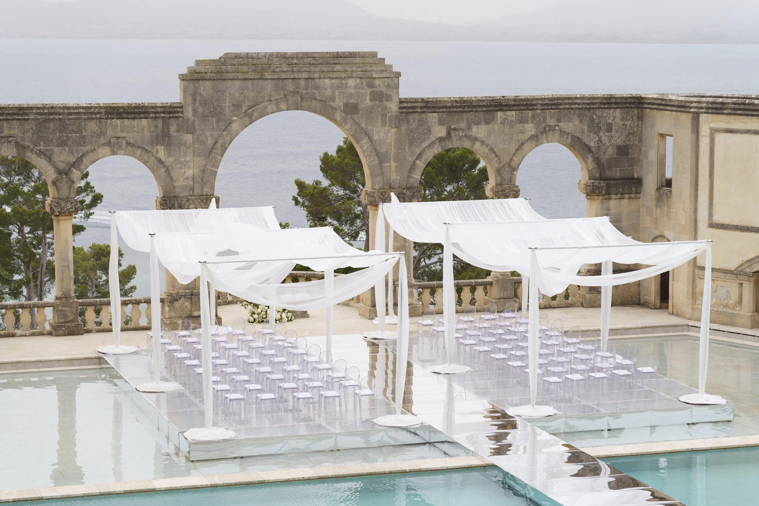 A wedding ceremony setup with chairs under white draped fabric, on a platform next to a swimming pool, with an ancient stone arch and view of the sea in the background.