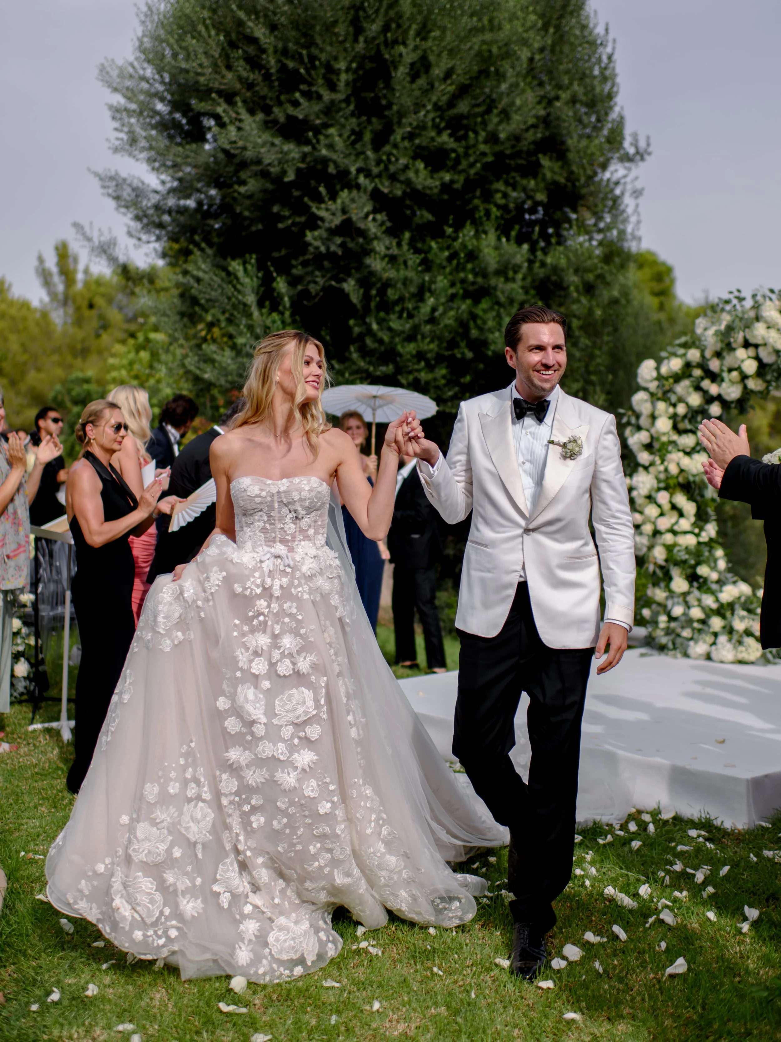 Bride and groom walking down outdoor wedding aisle, smiling, holding hands, guests clapping, large tree and floral arrangements in background.