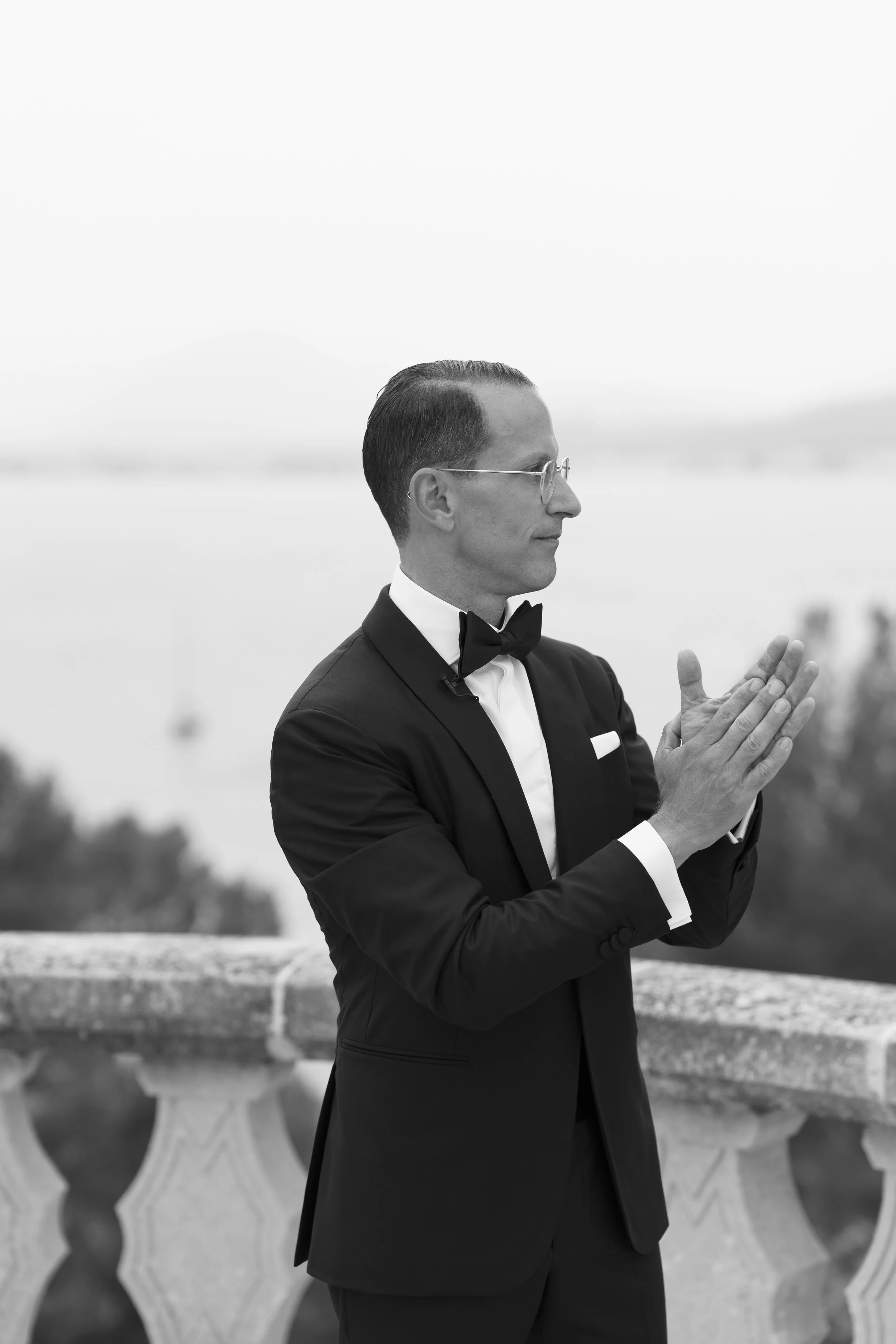 A man in a tuxedo with a bow tie and glasses, standing outdoors near a railing, with an out-of-focus body of water and trees in the background, captured in black and white.