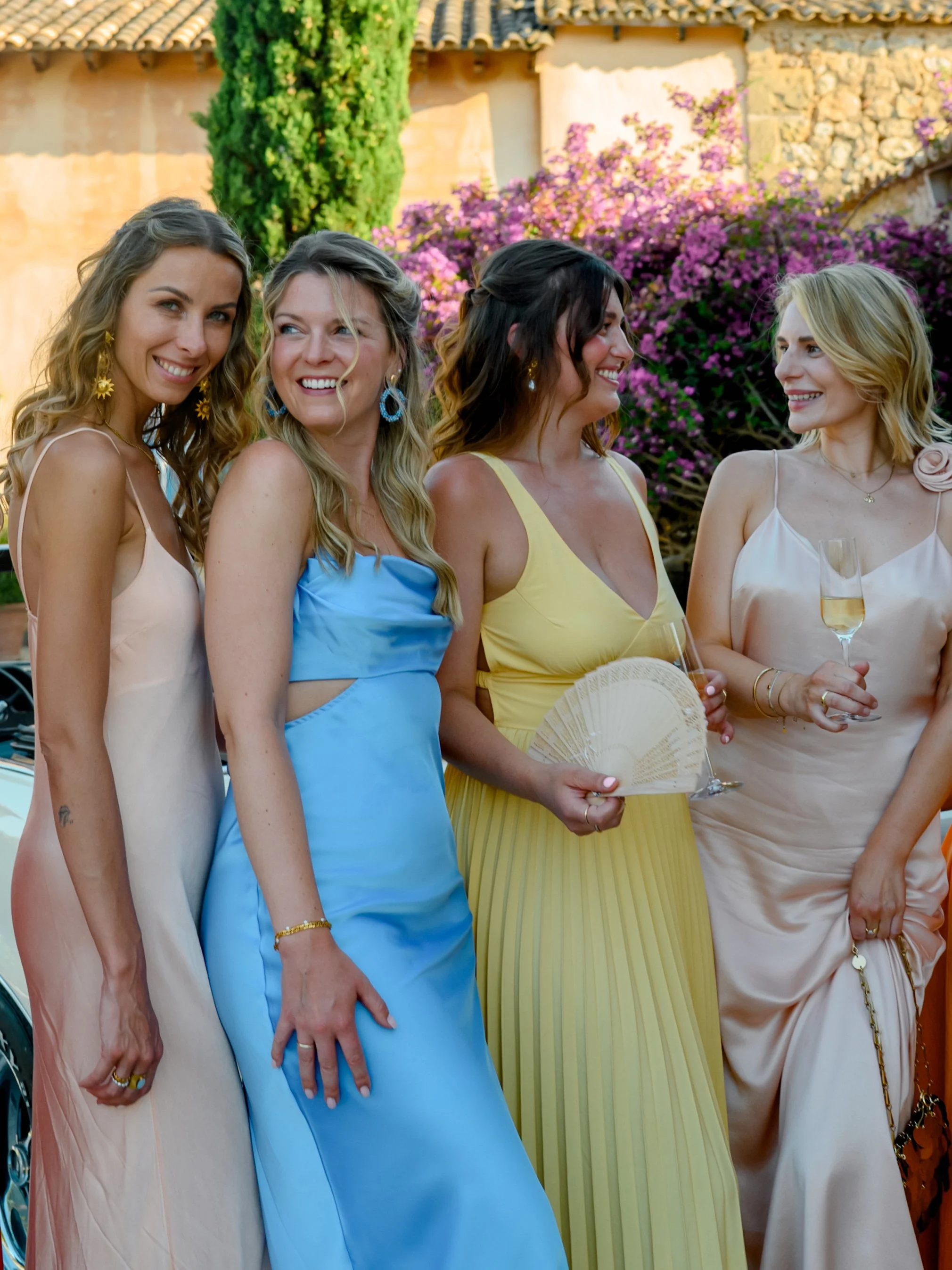 Four women dressed in colorful evening gowns standing together outdoors, holding glasses of champagne, smiling and chatting with a background of flowers and a beige building.