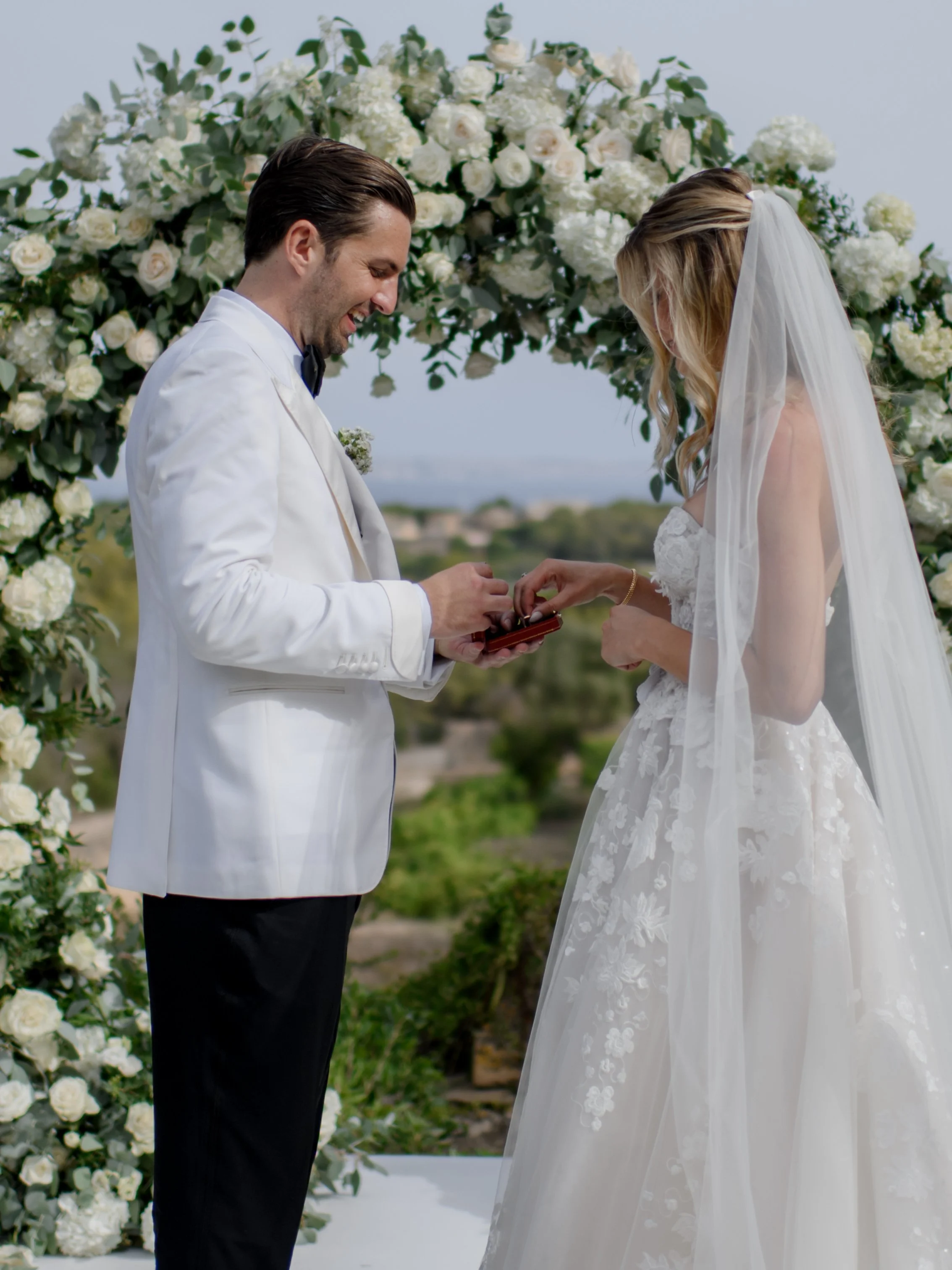 A bride and groom exchanging rings during their wedding ceremony outdoors under a floral arch.
