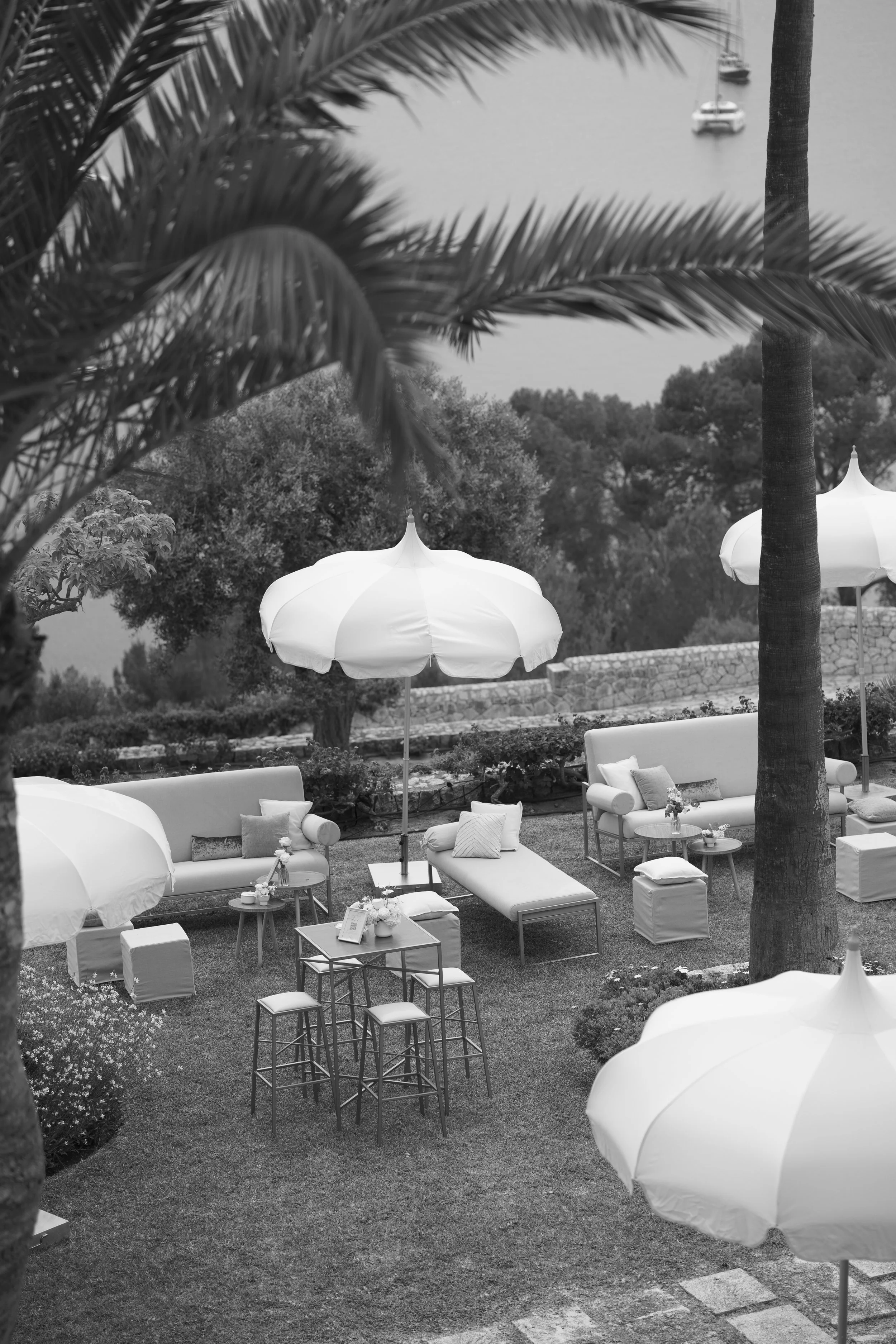 Outdoor seating area with white umbrellas, sofas, and chairs by a river with trees and a boat in the background.