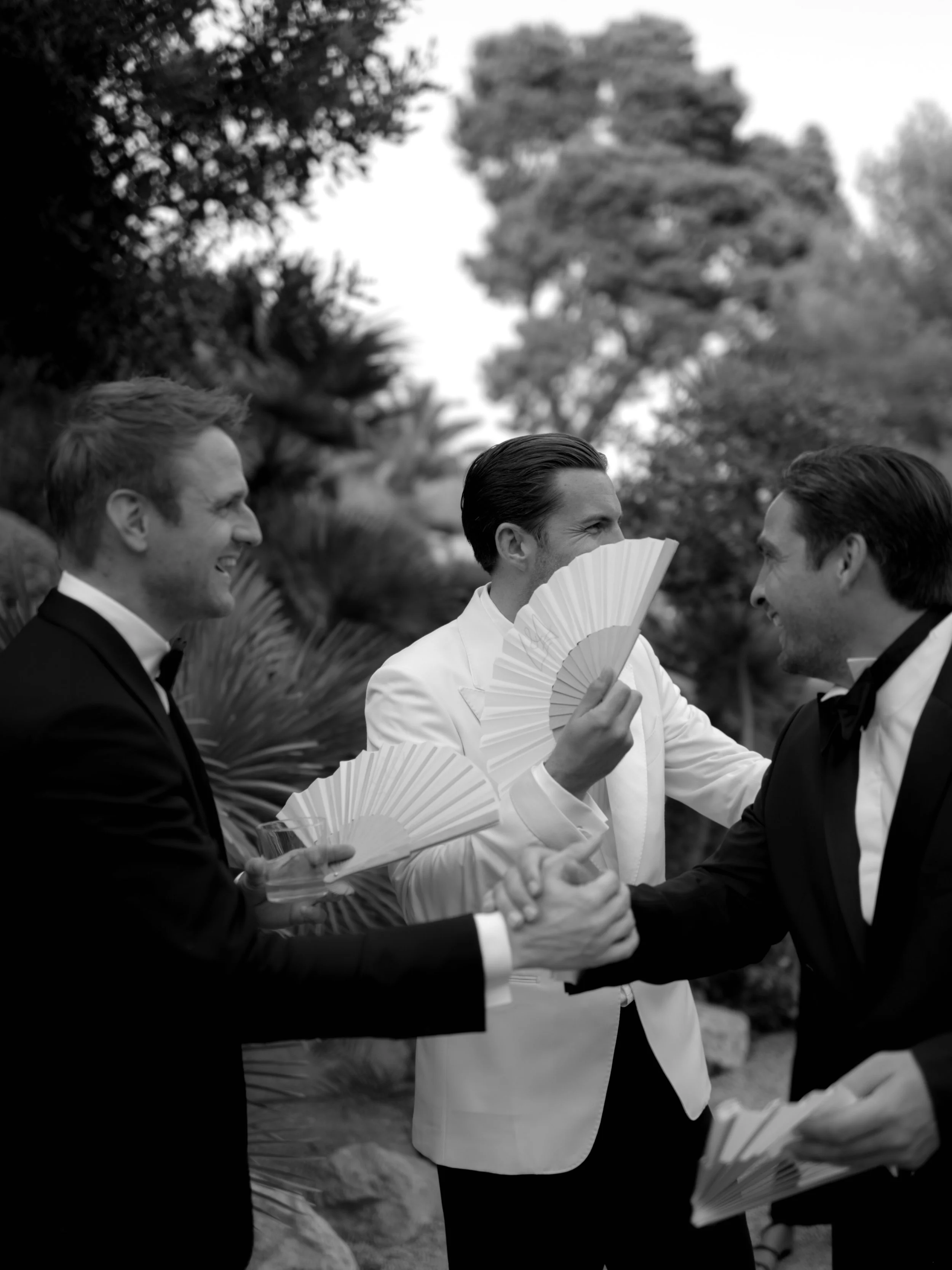 Three men in tuxedos at an outdoor event, engaging in conversation and handshakes, with trees in the background.
