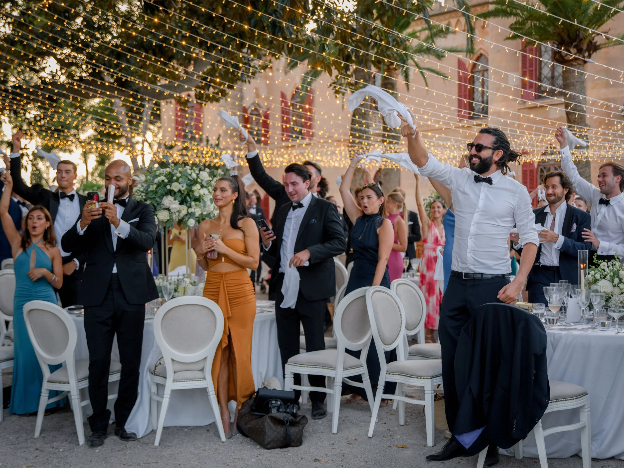 Guests at a formal outdoor event, possibly a wedding reception, dancing under string lights with trees and a building in the background.