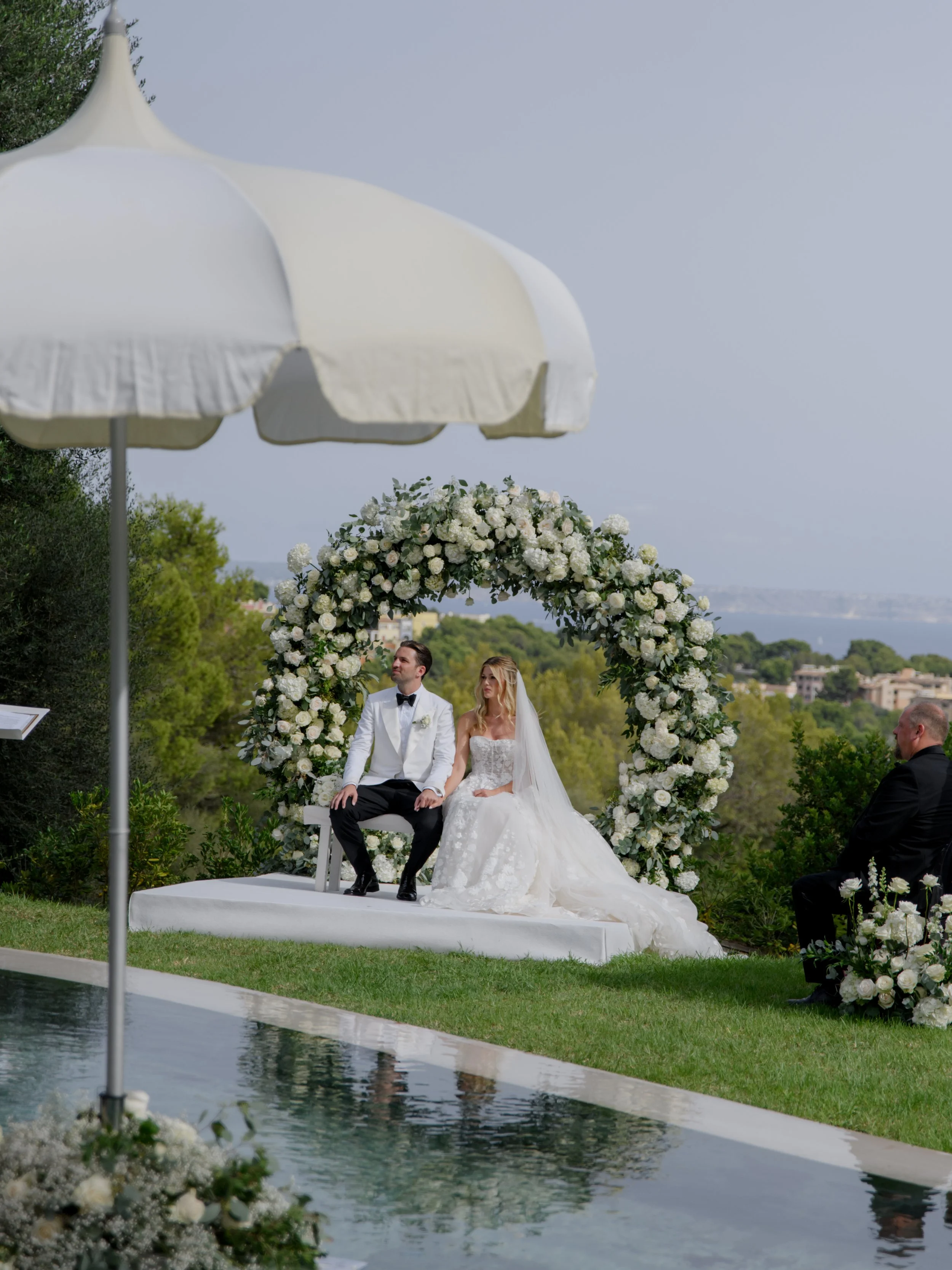 A bride and groom sitting on a white platform during an outdoor wedding ceremony, with a large floral arch behind them, overlooking a scenic landscape with trees and water in the background.