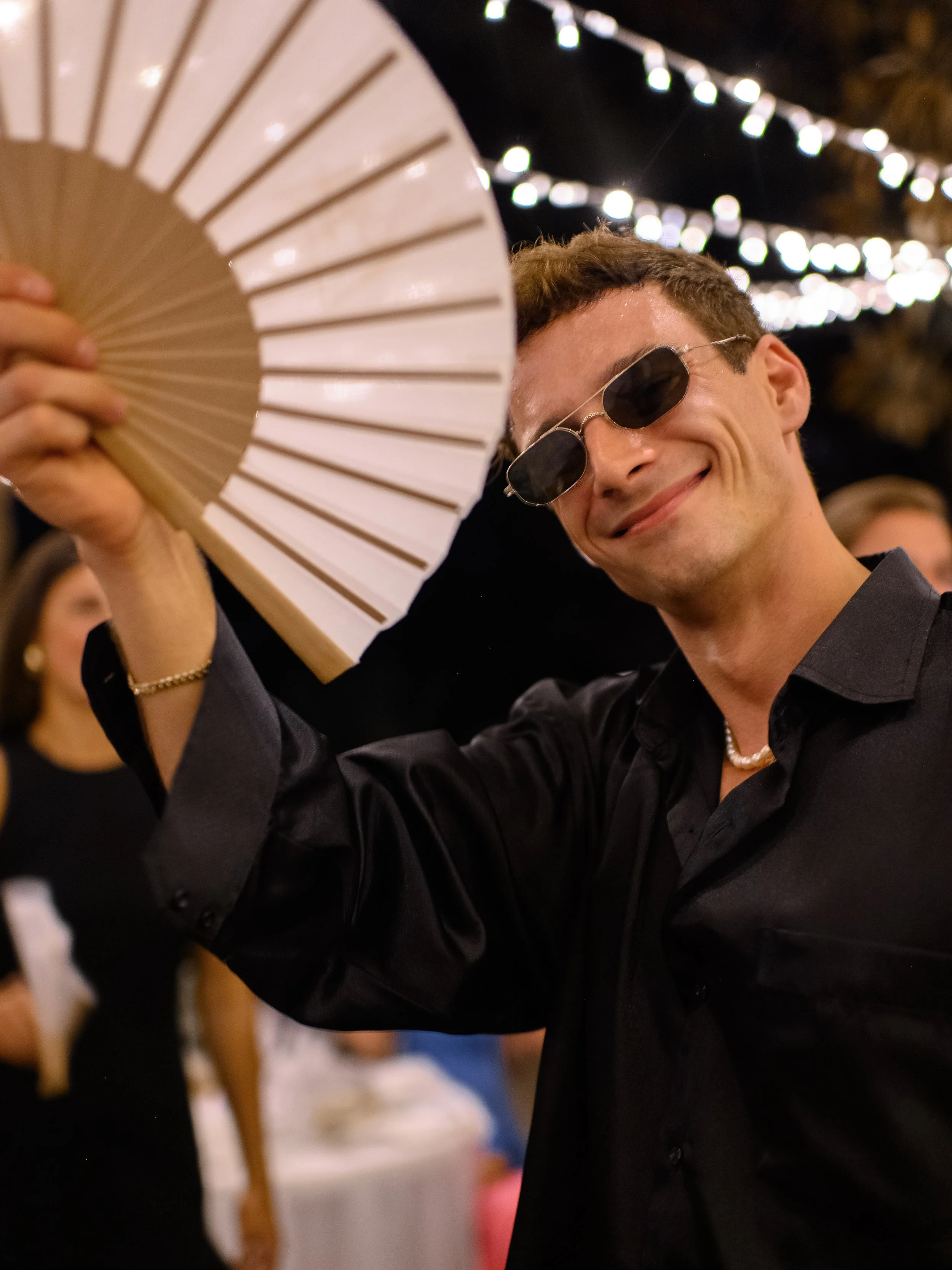 A young man smiling while holding a paper fan at an outdoor event with string lights.