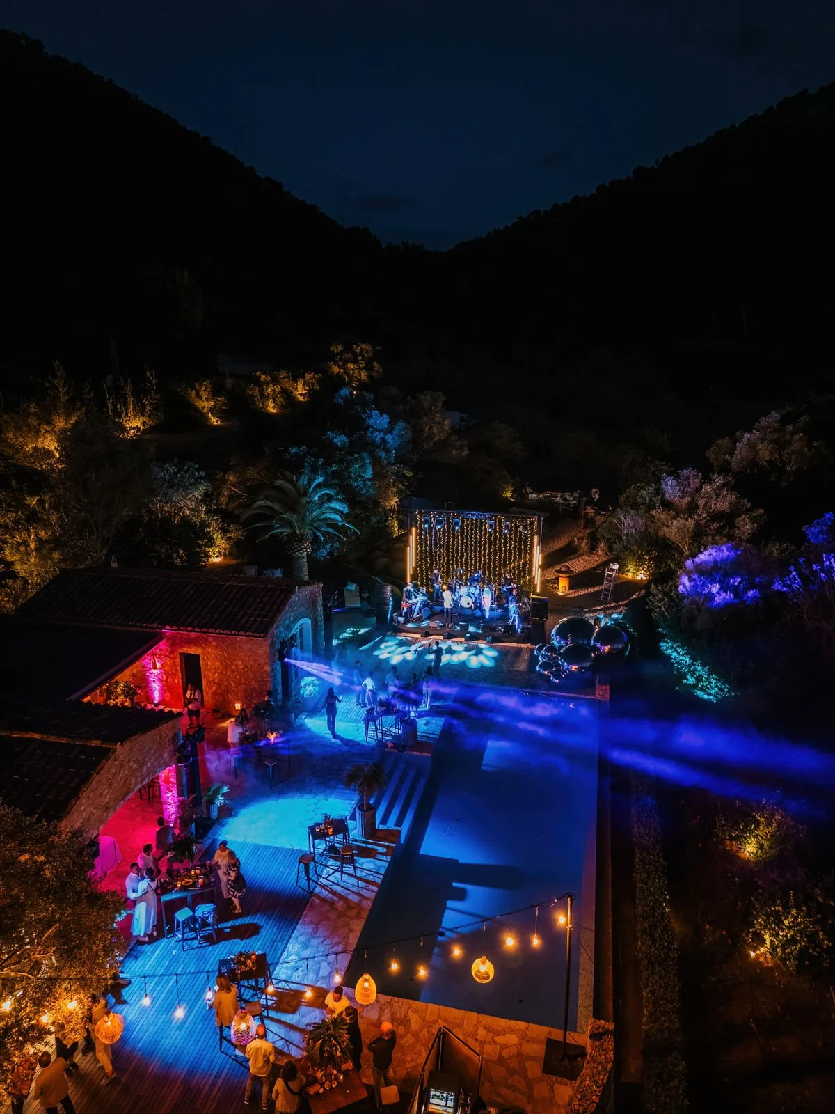 Nighttime outdoor party with a stage, musical band, colorful lighting, tables, and people socializing.