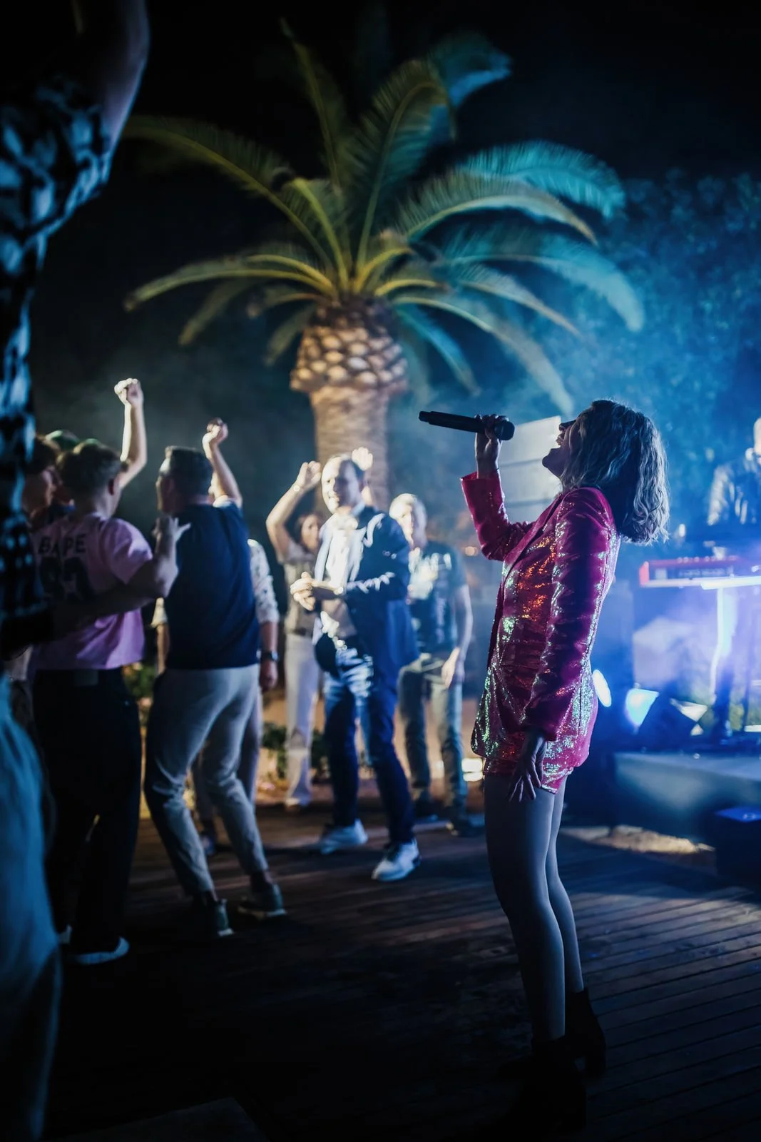 A woman in a shiny red jacket singing into a microphone at a lively outdoor party at night with palm trees in the background.