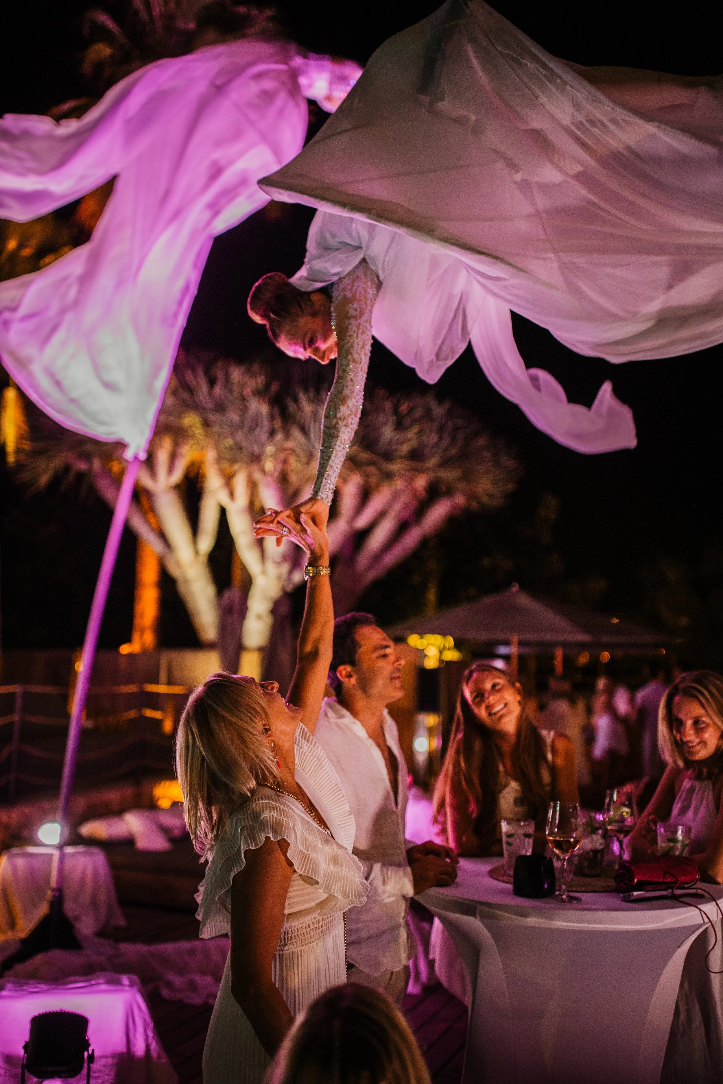 People at an outdoor night event watching a performer in a flowing costume suspended above them.