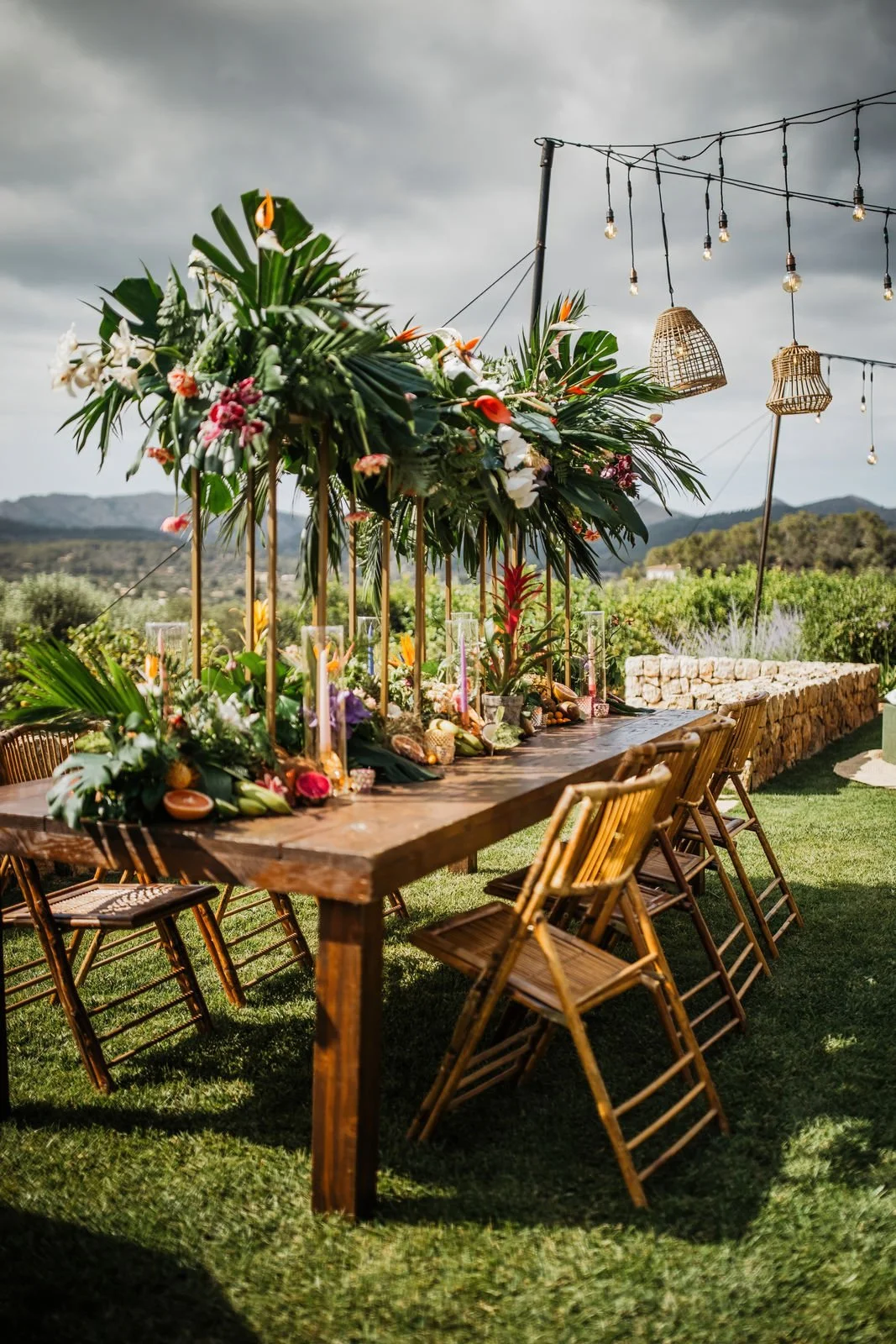 Outdoor dining table decorated with tropical flowers and greenery, surrounded by wooden chairs, under string lights, against a backdrop of rolling hills and a cloudy sky.