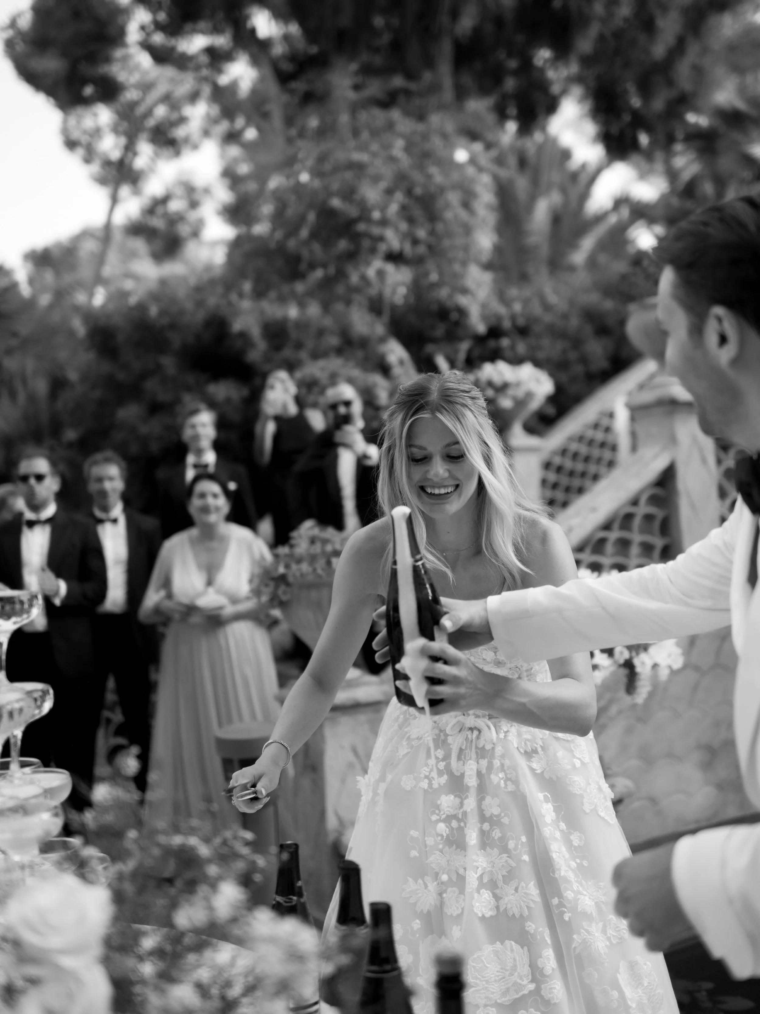 A woman in a wedding dress is smiling as a man in a tuxedo hands her a bottle of champagne at an outdoor wedding reception. Several guests are in the background, some in formal attire, standing in a garden setting.