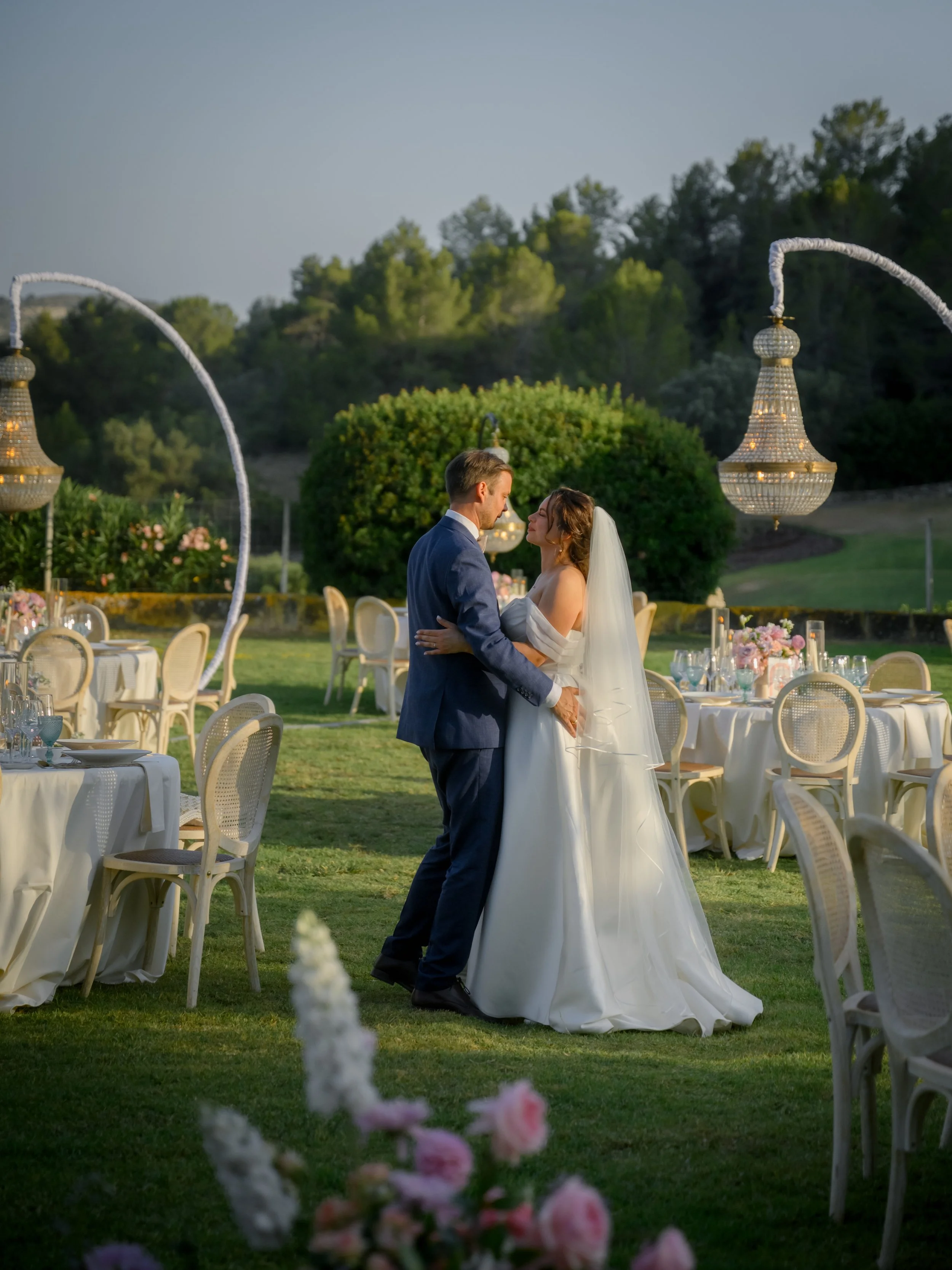 A bride and groom share a dance outdoors at their wedding reception in a garden, surrounded by decorated tables and chandeliers, with trees and a hill in the background.