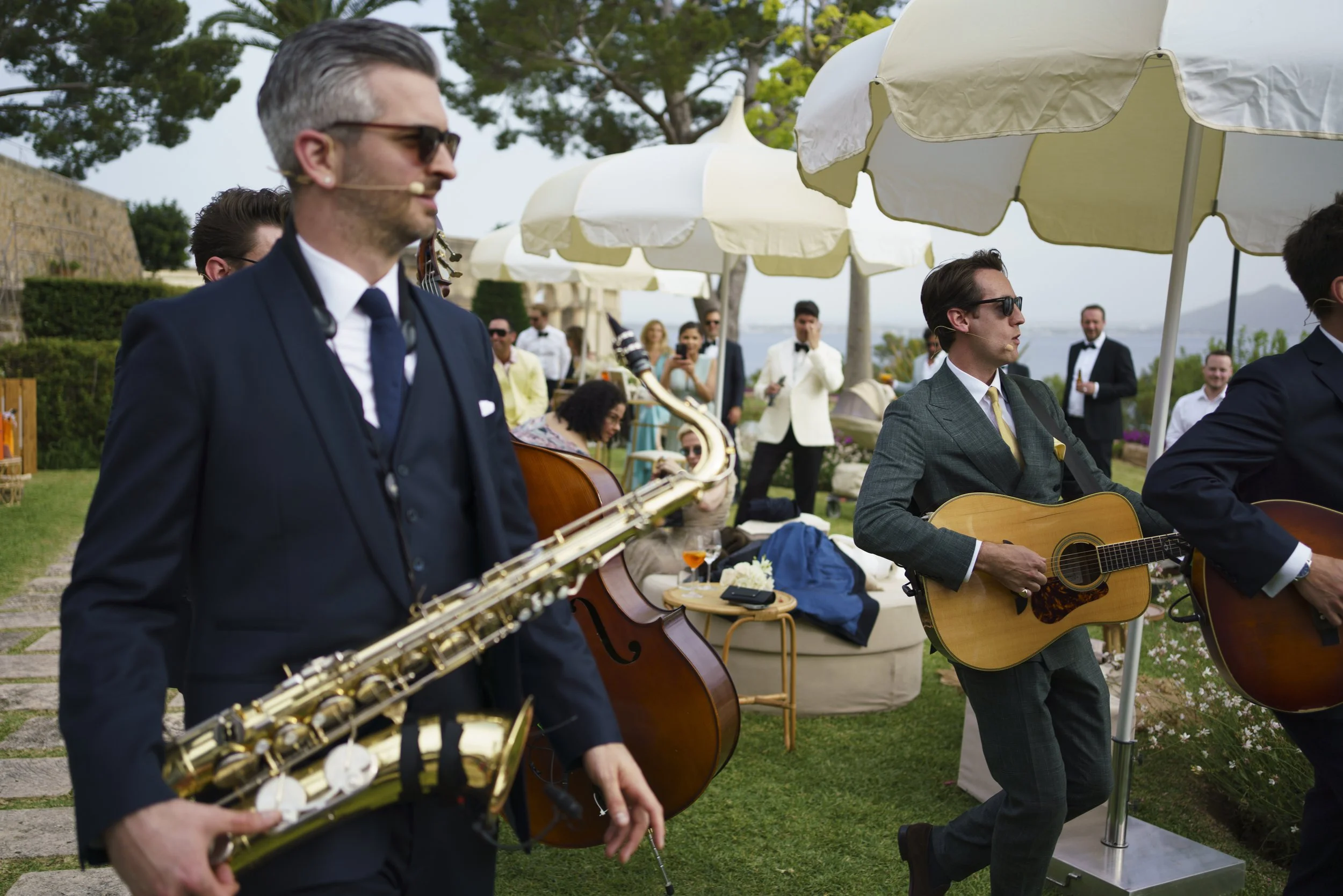 Musicians performing at an outdoor event, with one playing a saxophone and another a guitar, under umbrellas with a gathering of people in the background.