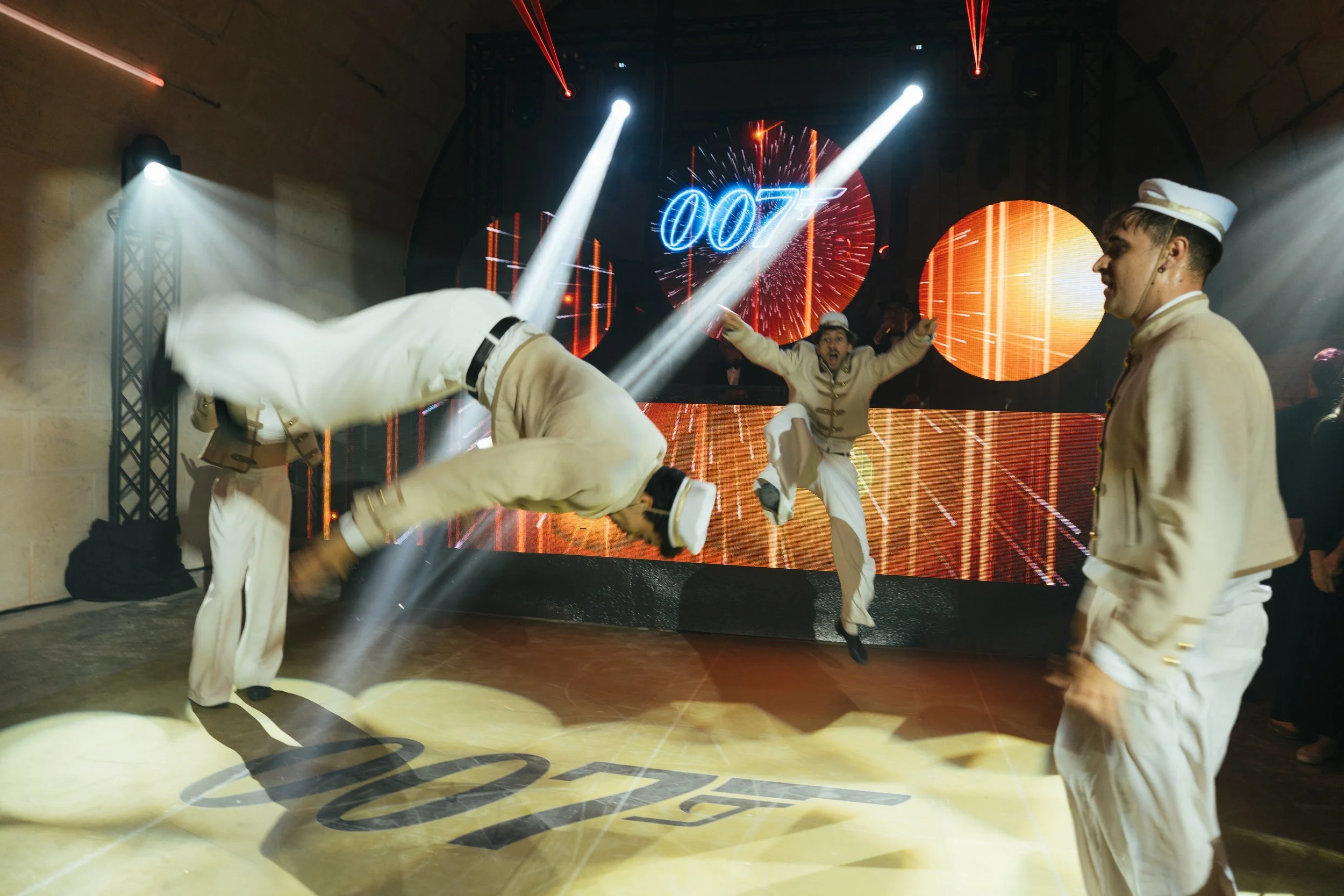 Dancers dressed in 007-themed costumes performing on a dance floor with colorful lights and a large screen displaying '007' in the background.