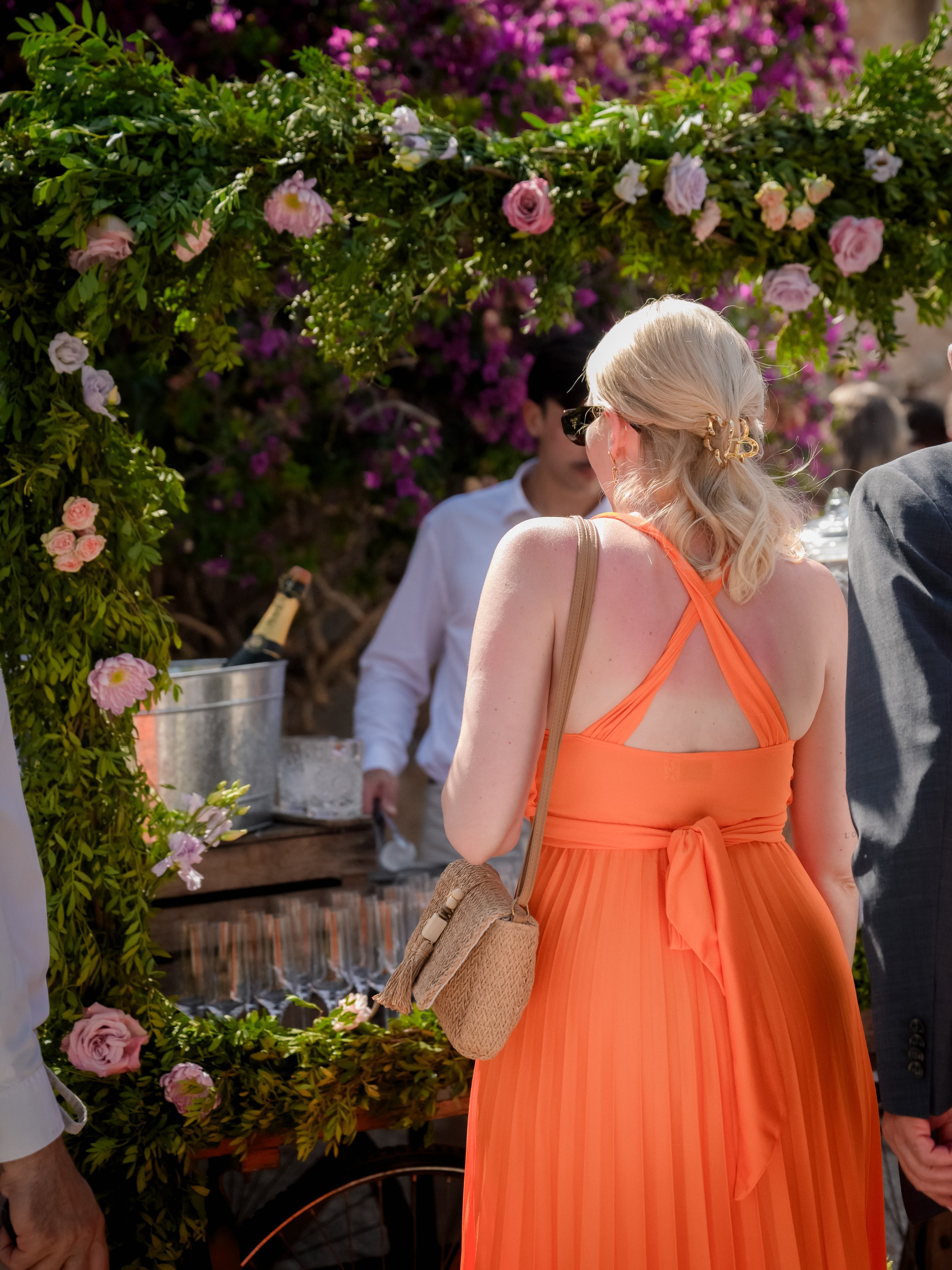 A woman with blonde hair in a low ponytail wearing sunglasses and an orange dress stands at a floral wedding arch decorated with pink and purple flowers. A person in a white shirt is visible behind her, with a champagne bottle in an ice bucket nearby