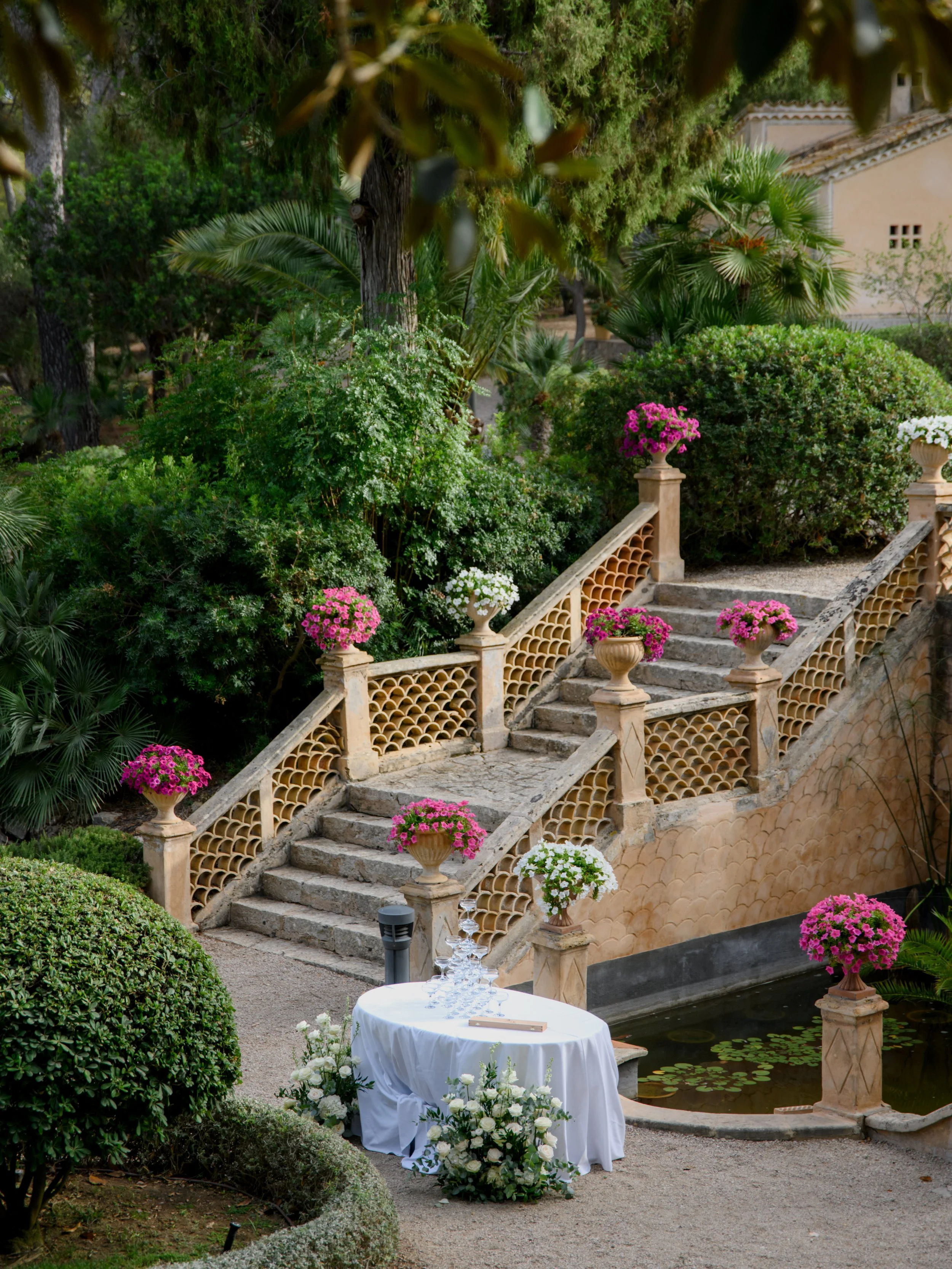 A garden scene with stone stairs decorated with pink and white flowering pots, a small pond with water lilies, a table with glasses and floral arrangements, surrounded by lush green trees and shrubs.