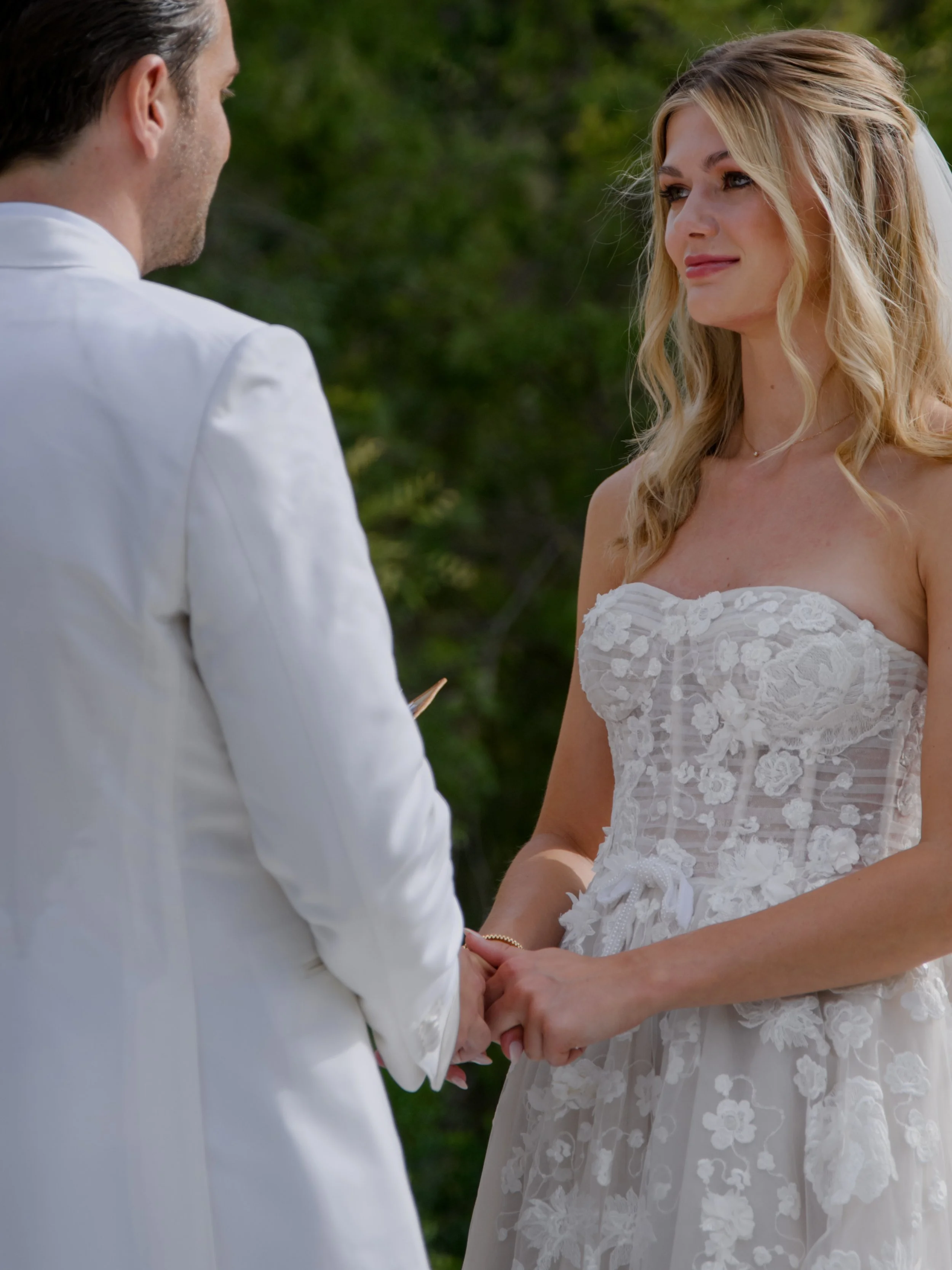 A bride and groom holding hands during their wedding ceremony outdoors, with the bride in a lace wedding dress and the groom in a white suit.