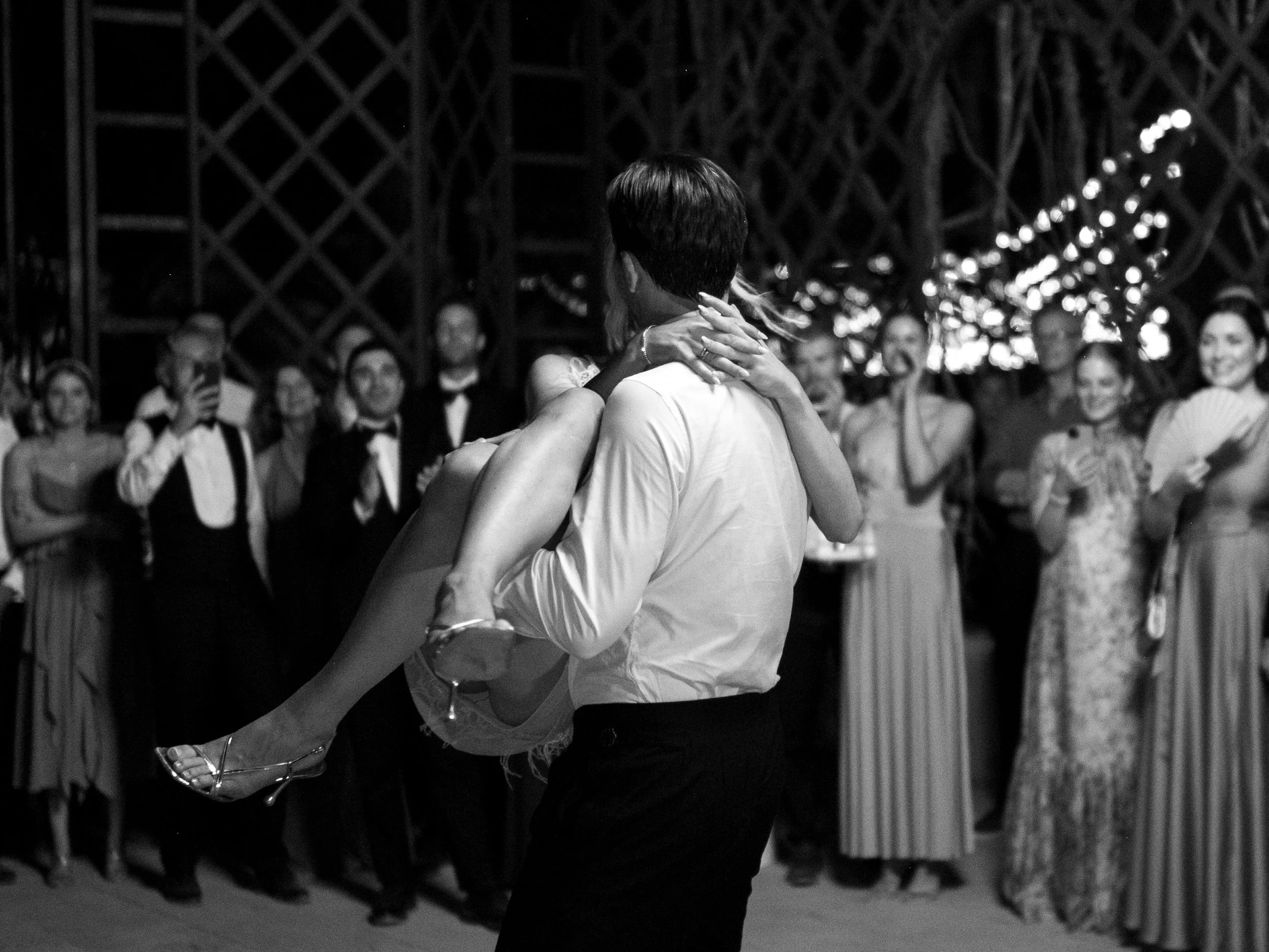 A man in a white shirt and black pants is dancing with a woman in a wedding dress at a wedding reception, surrounded by guests in formal attire watching and smiling.