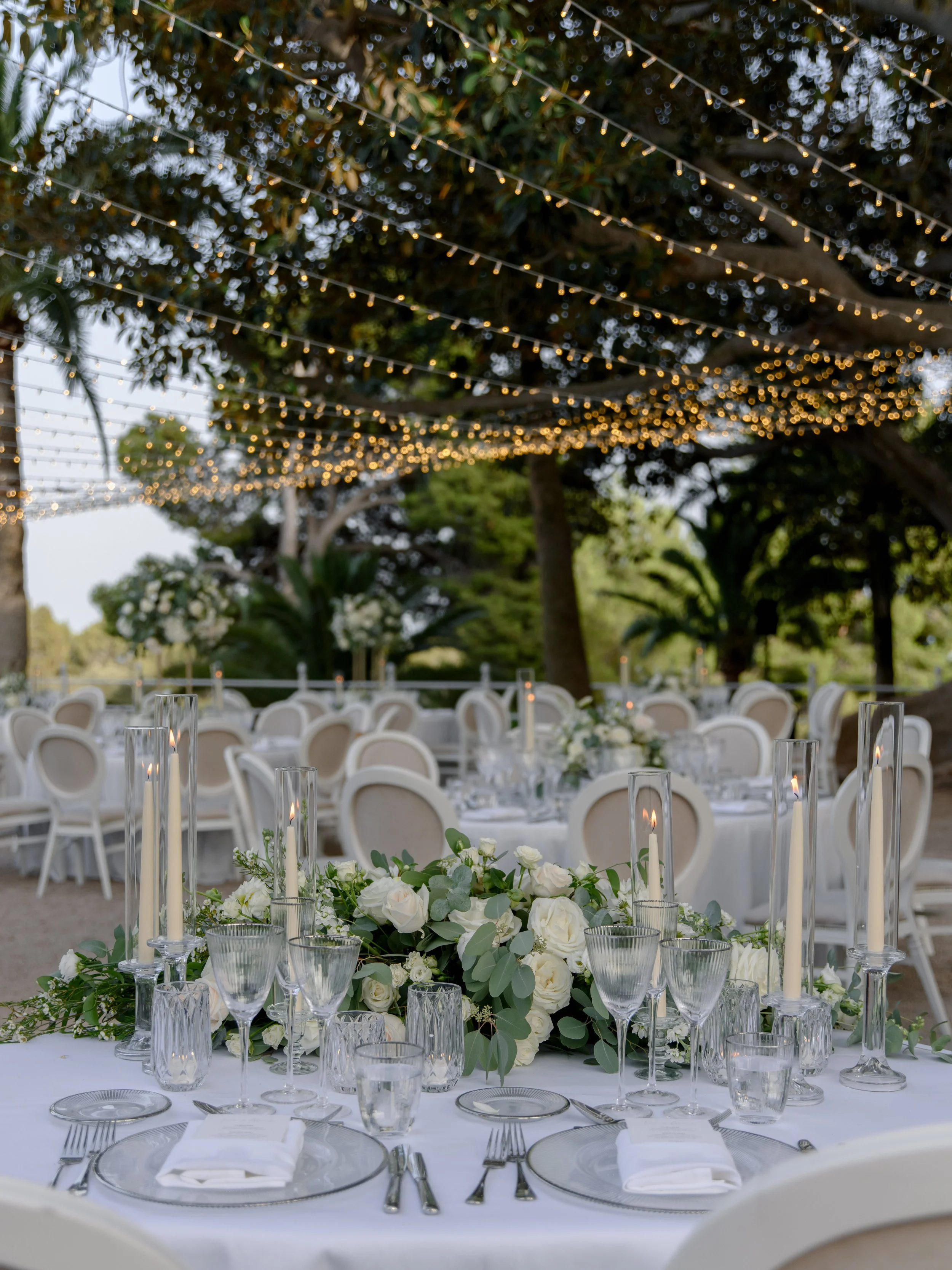 Elegant outdoor event setup with round tables, white chairs, floral centerpieces, and string lights hanging from trees.